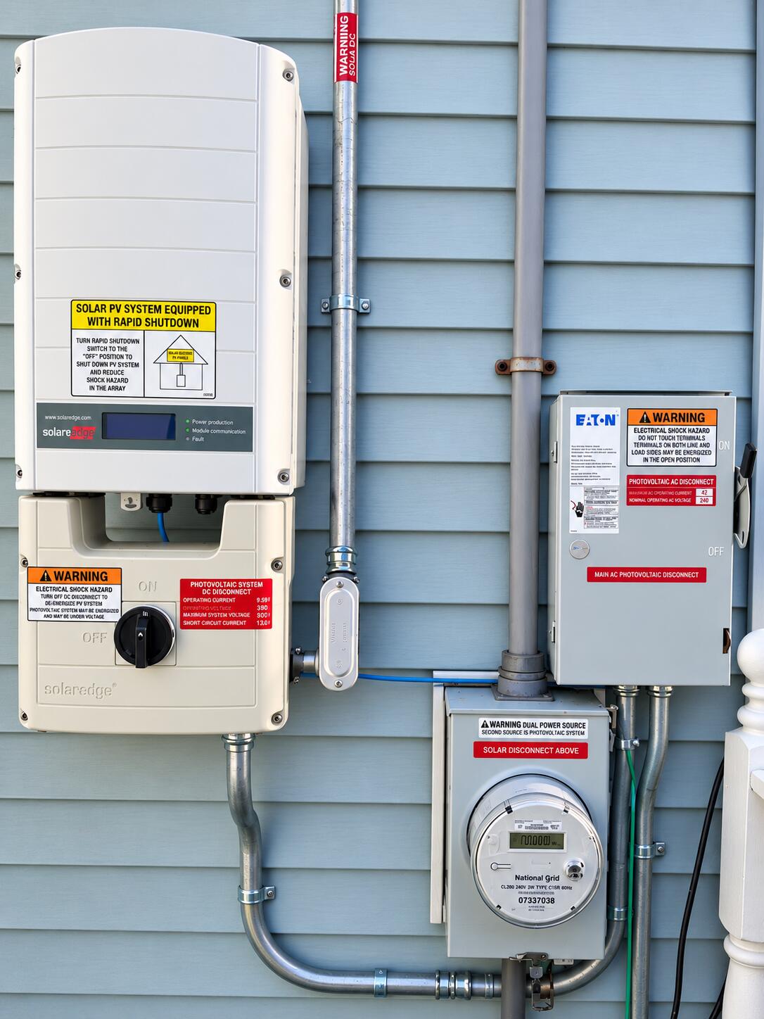 Solar power equipment on blue siding, including a SolarEdge inverter, disconnect switches, and electric meter.
