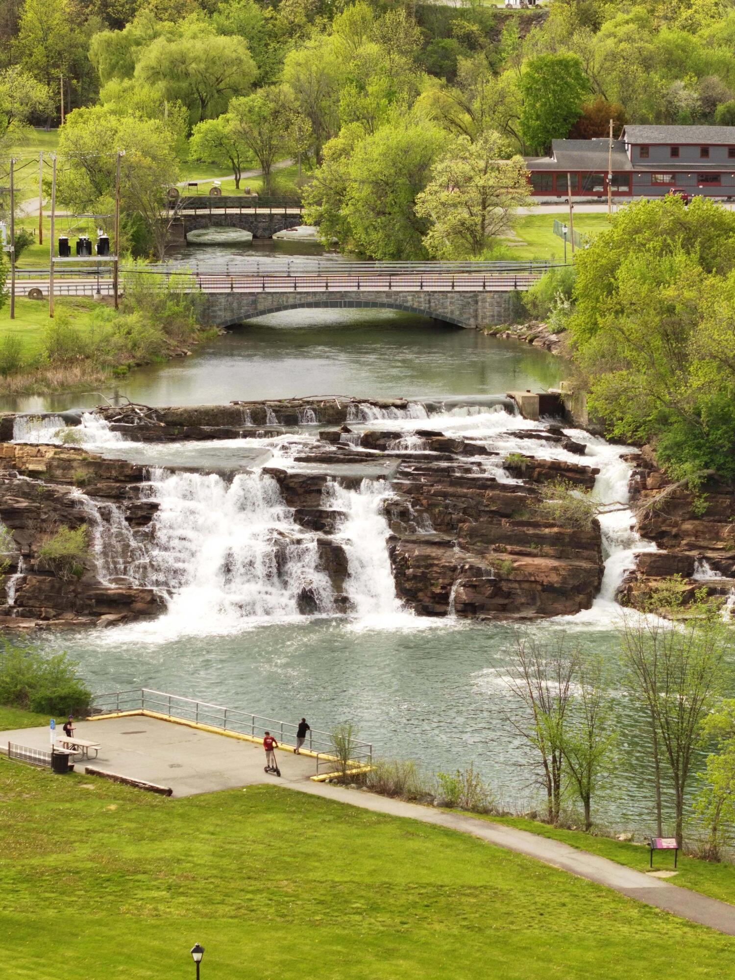 River waterfall below two stone bridges, with a park viewing area and green trees.