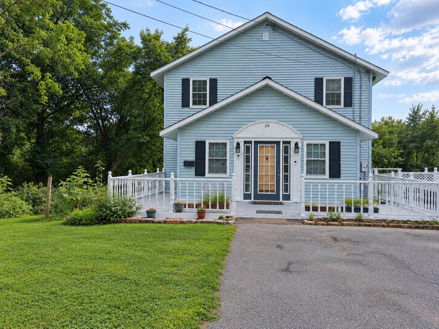 Two-story light blue house with white railings, black shutters, and a paved driveway.