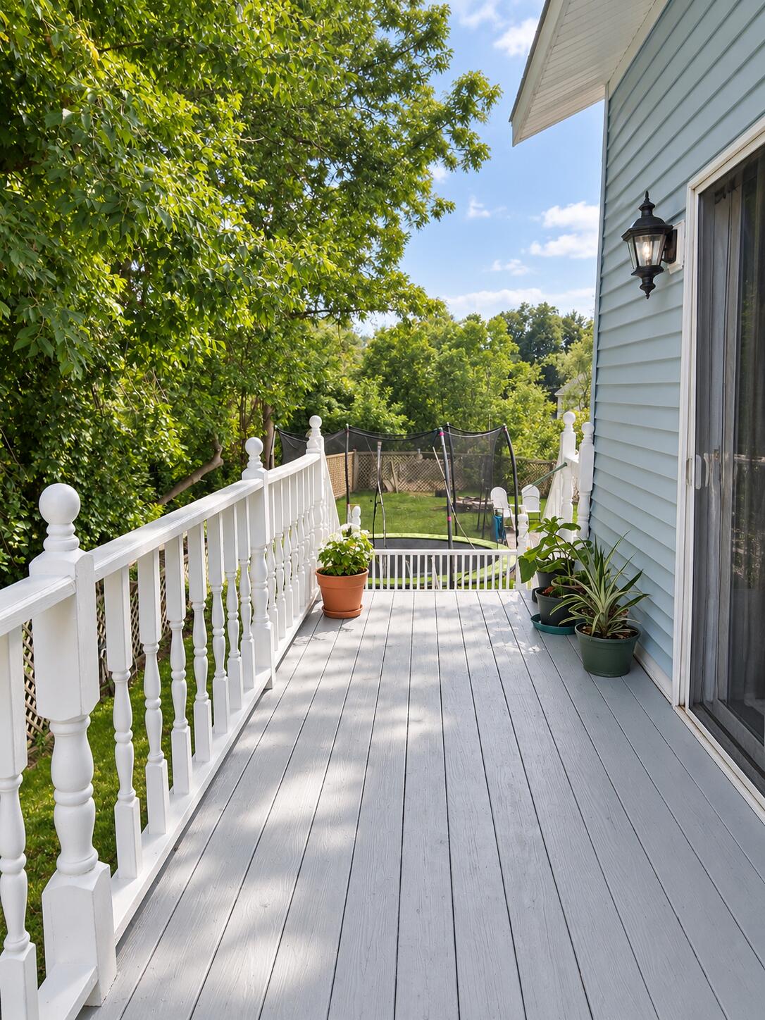 Gray deck with white railings beside a light blue house, overlooking a leafy yard and trampoline.