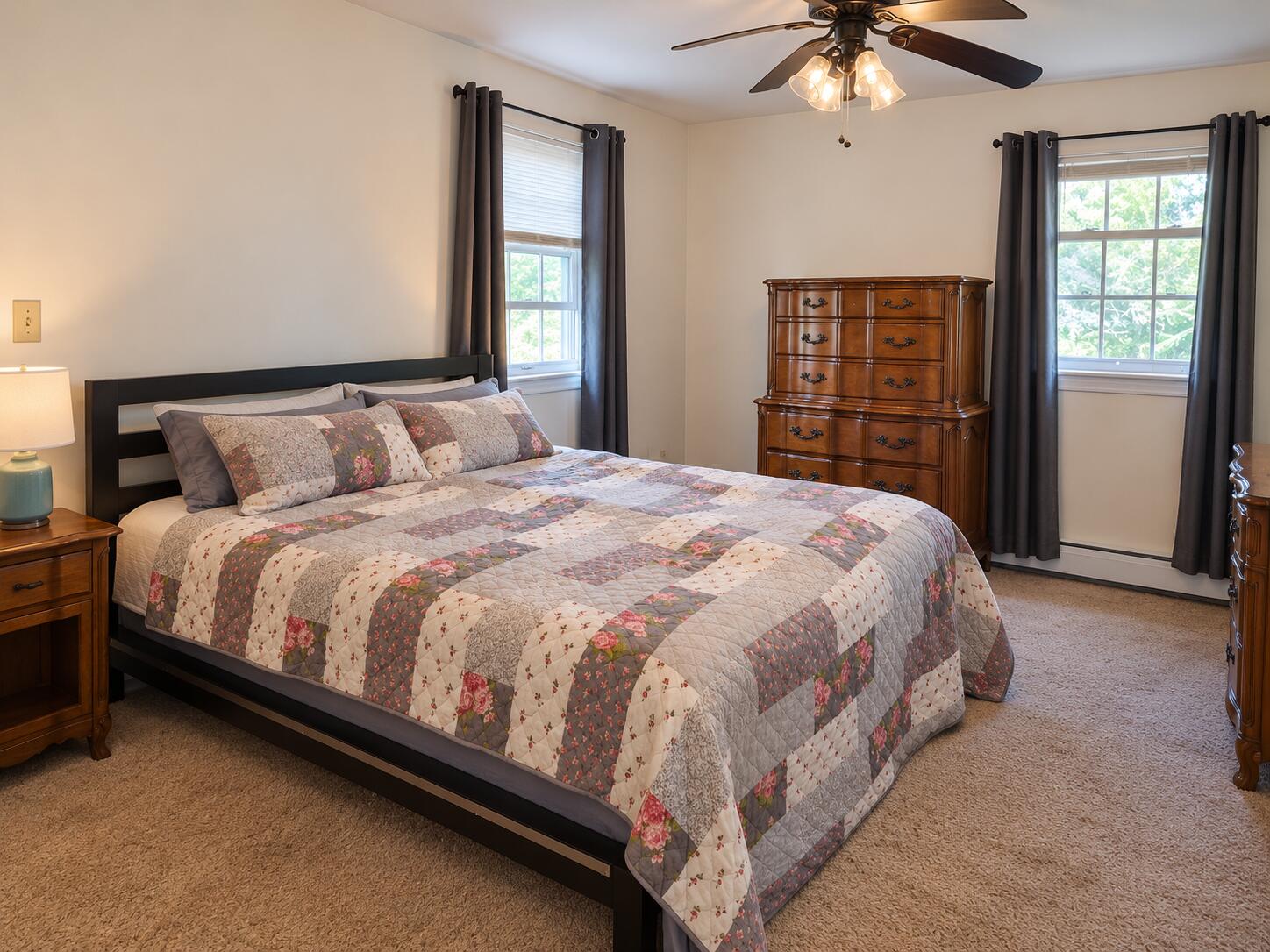 Bedroom with a floral patchwork quilt, wooden dressers, and two curtained windows.