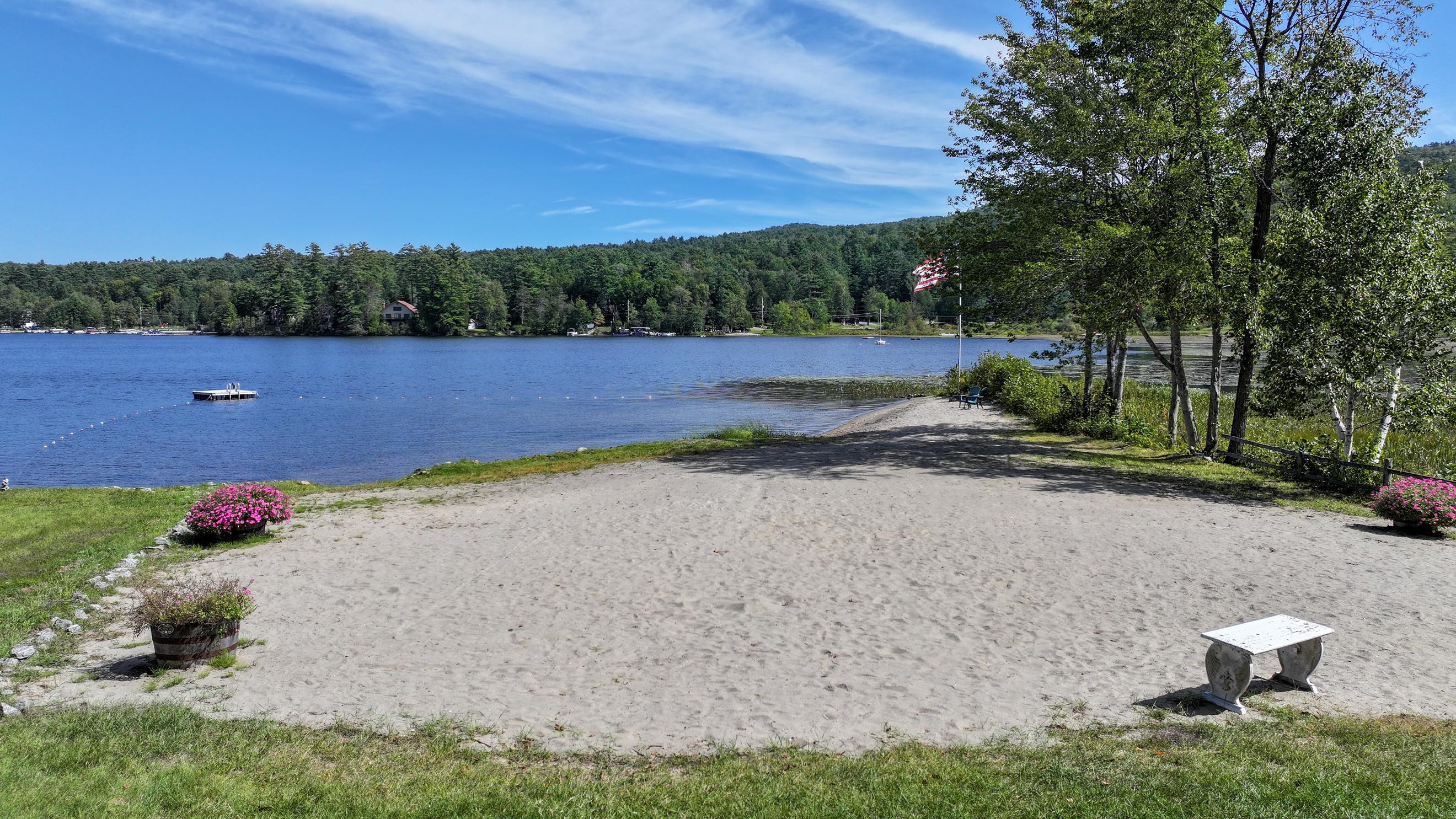 Sandy lakeside beach with a buoy-marked swimming area and wooded hills across the water.