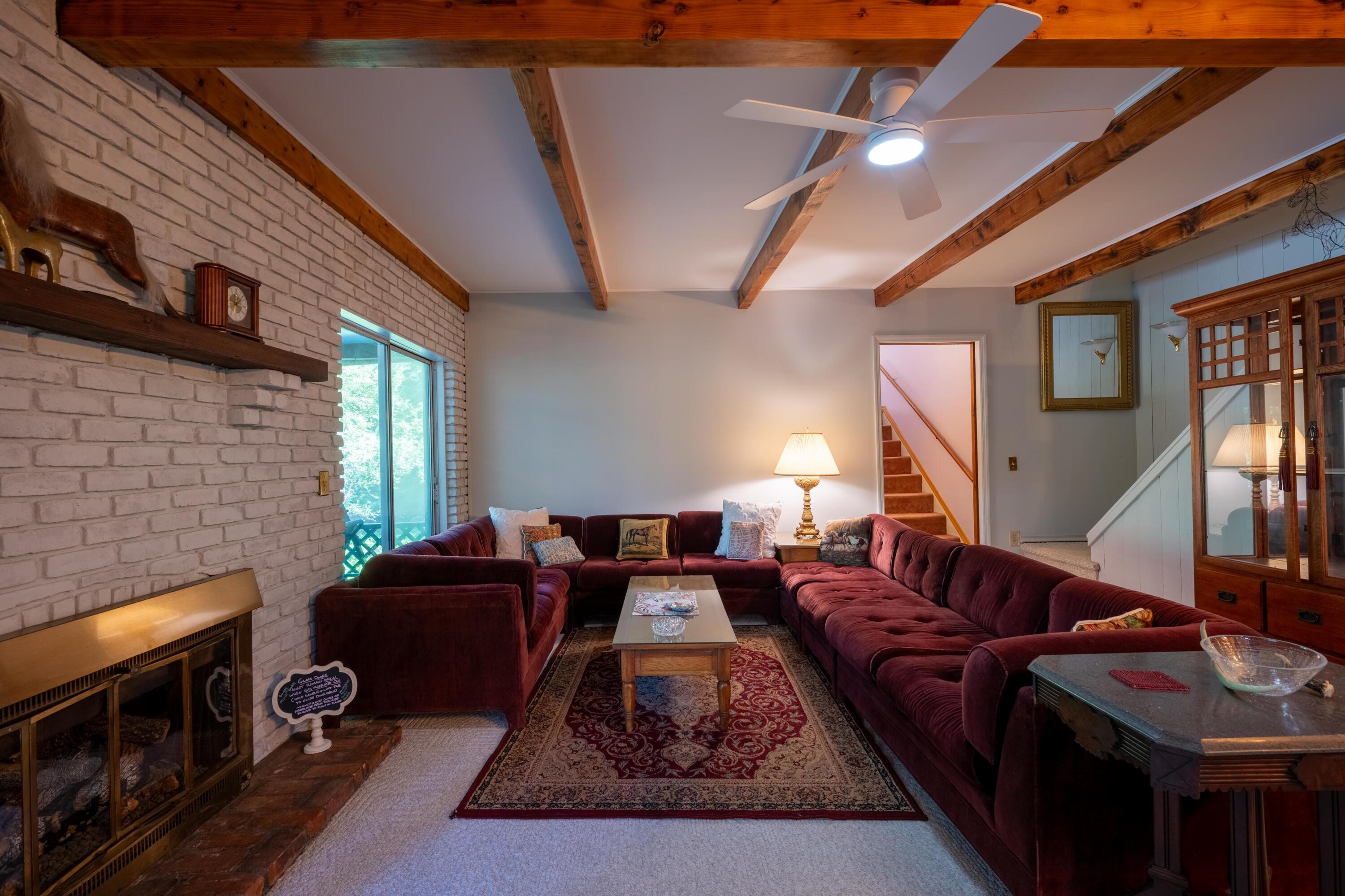 Living room with a burgundy sectional, white brick fireplace, and exposed wood ceiling beams.