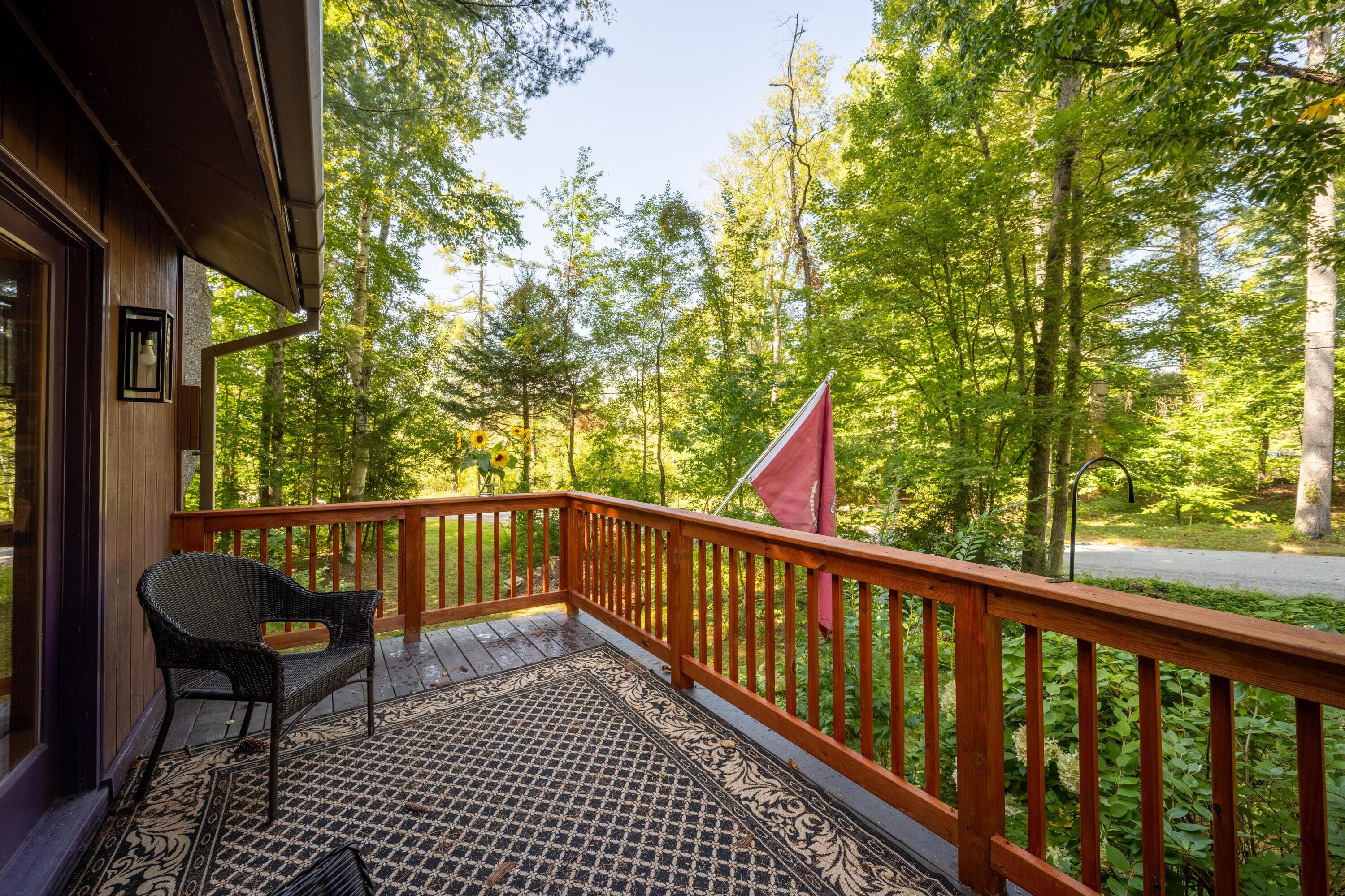 Wooden porch with a wicker chair and patterned rug overlooking a leafy wooded yard.