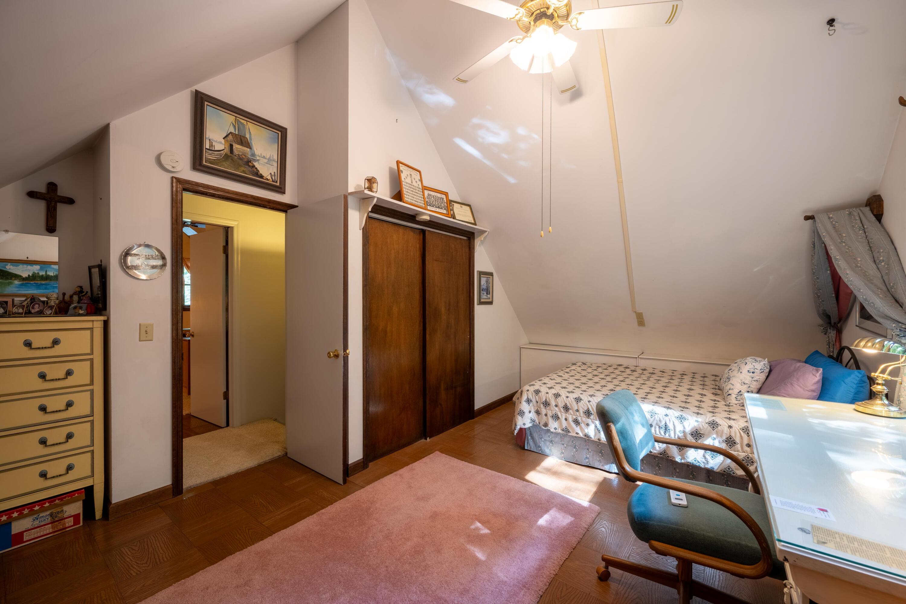 Attic bedroom with a sloped ceiling, patterned bed, wood closet doors, and sunlight across a pink rug.