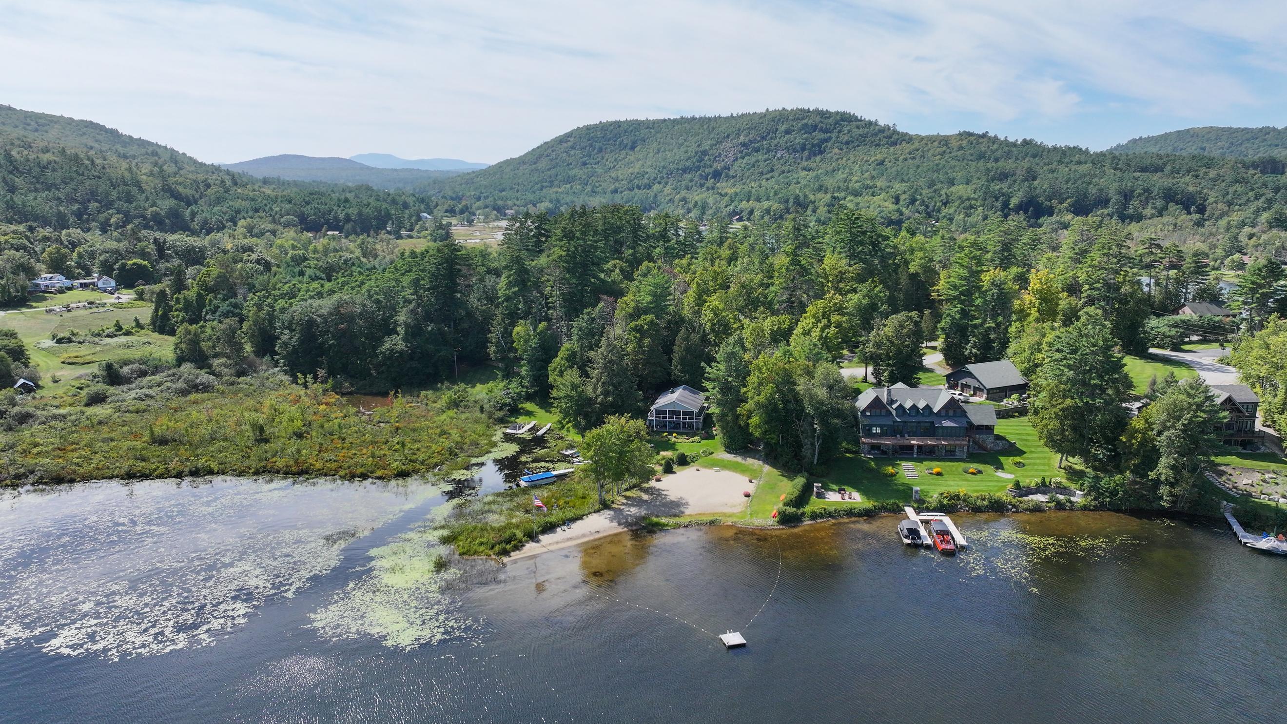 Aerial view of a lakeside home with a sandy beach, docks, and forested mountain views.