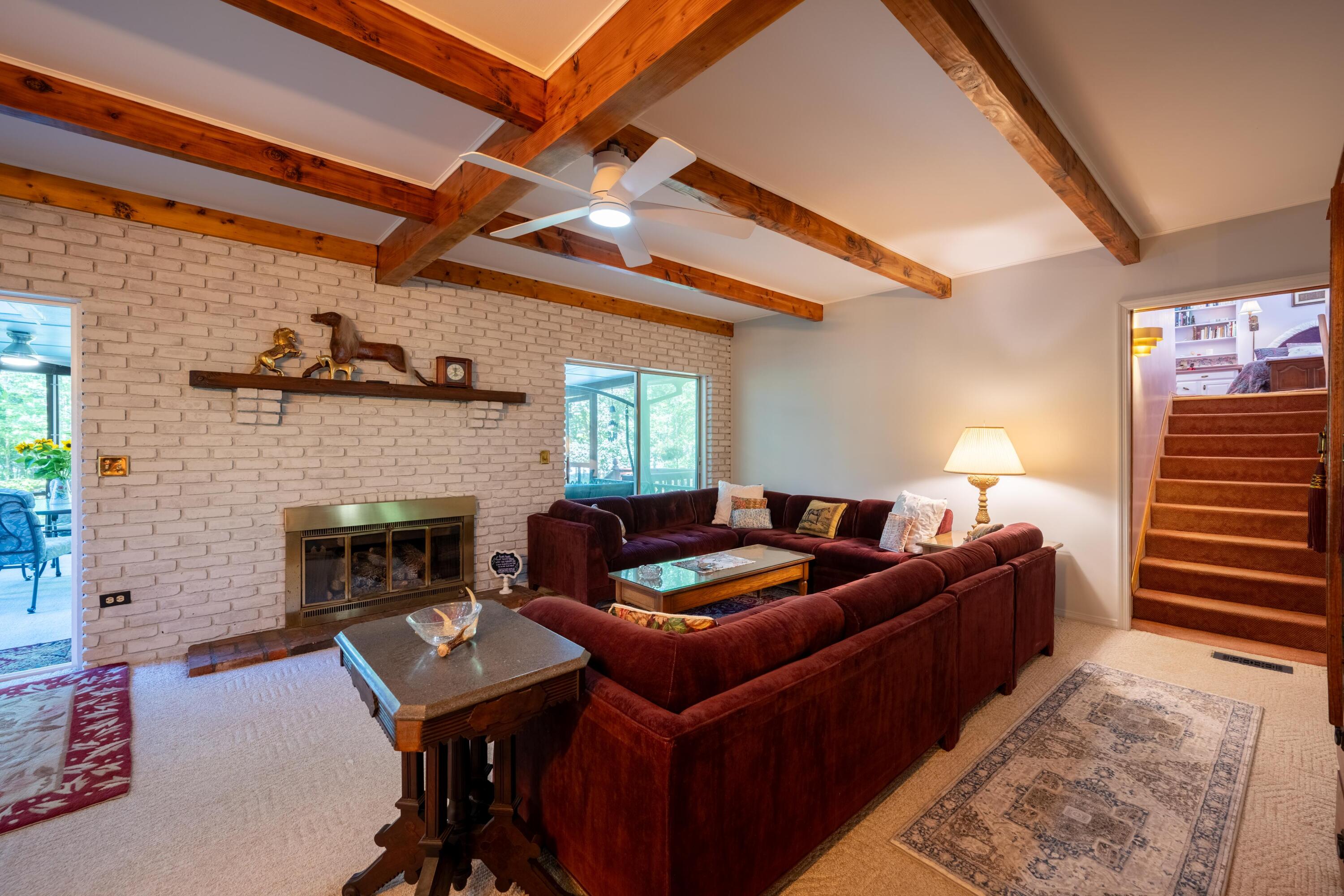 Living room with exposed ceiling beams, a white brick fireplace, burgundy sectional sofas, and carpeted stairs.