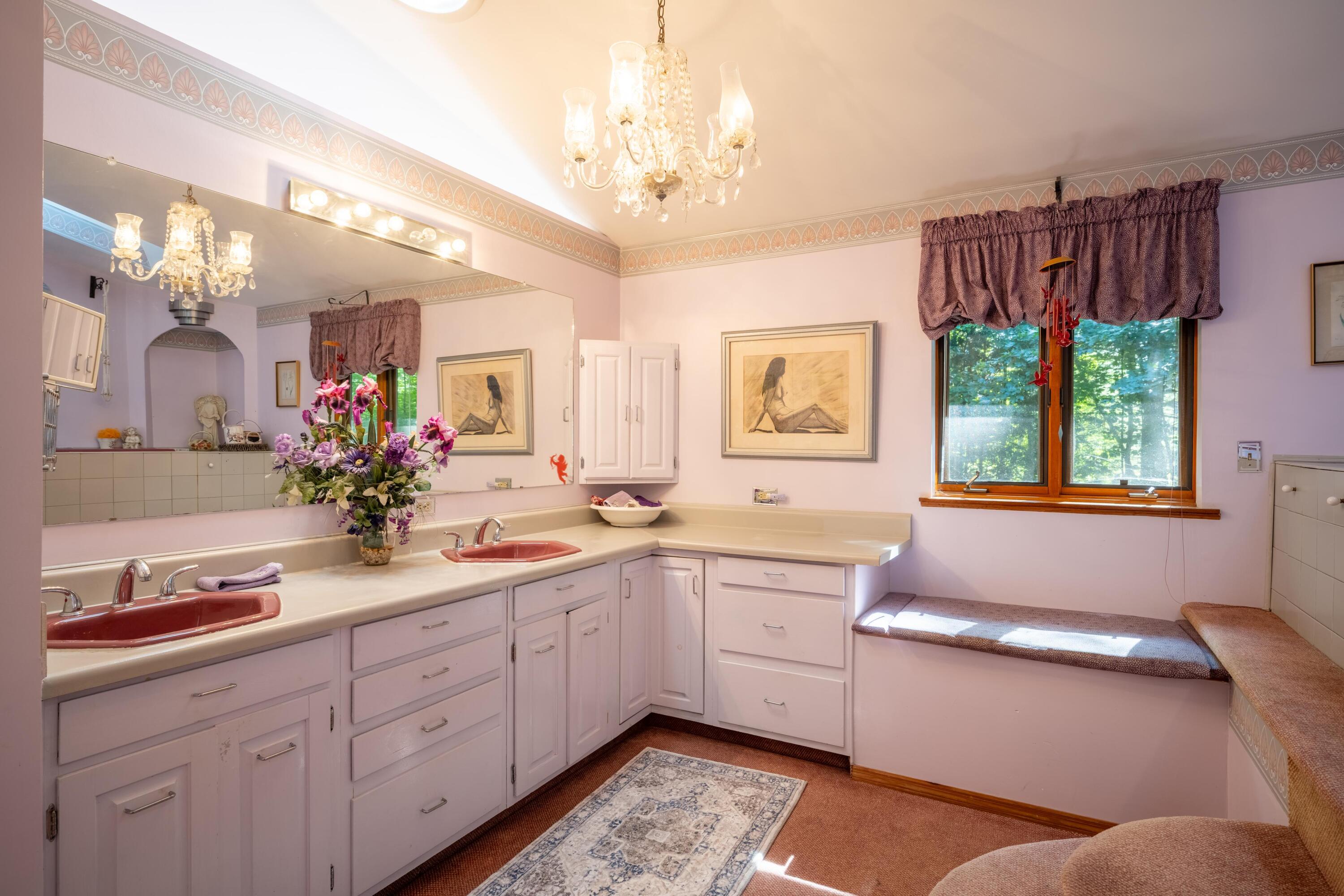 Bathroom with a double-sink vanity, chandelier, and built-in window seat overlooking greenery.