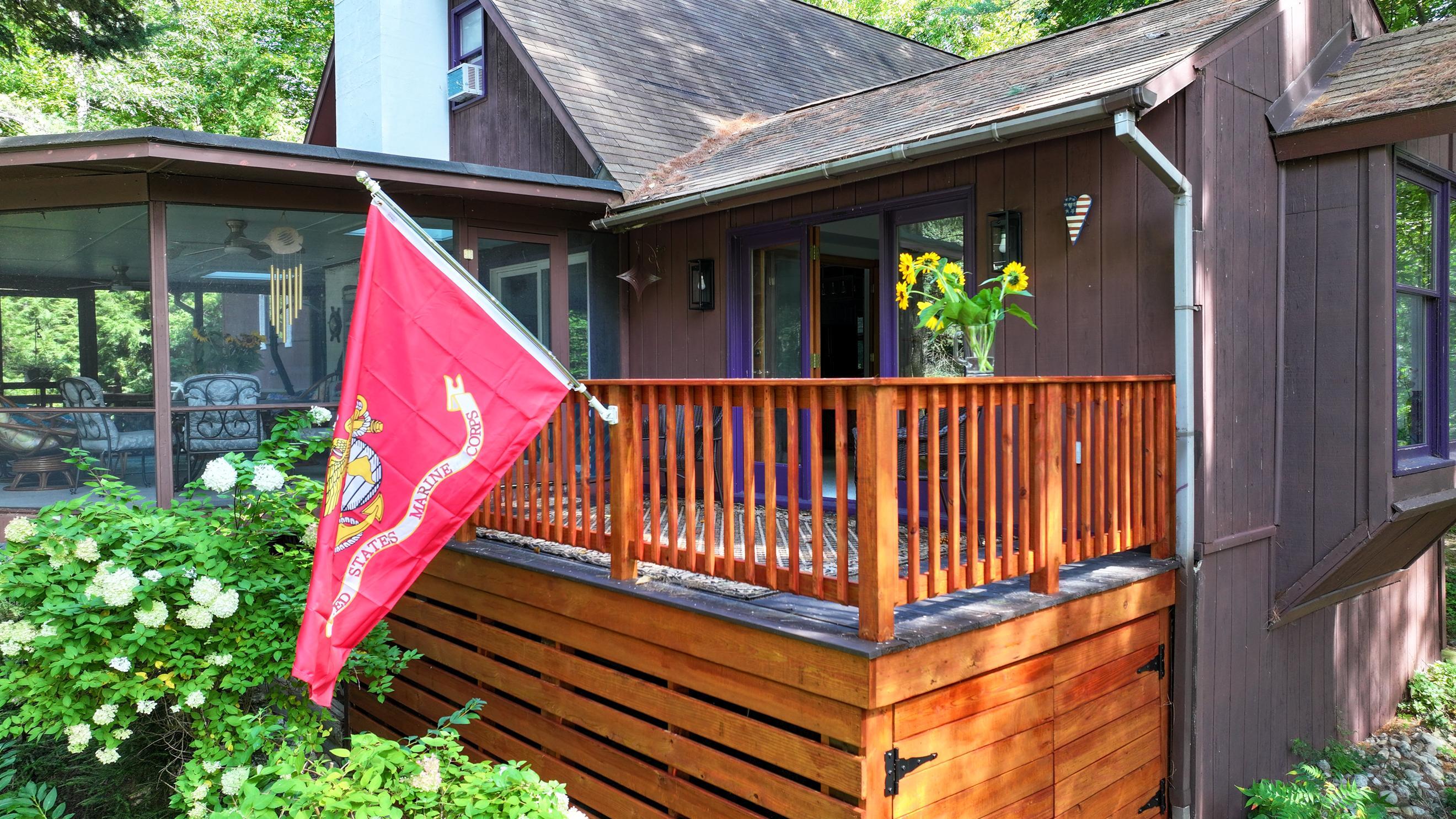 Brown-sided home with a raised wooden deck, screened porch, Marine Corps flag, and sunflowers by the entry.