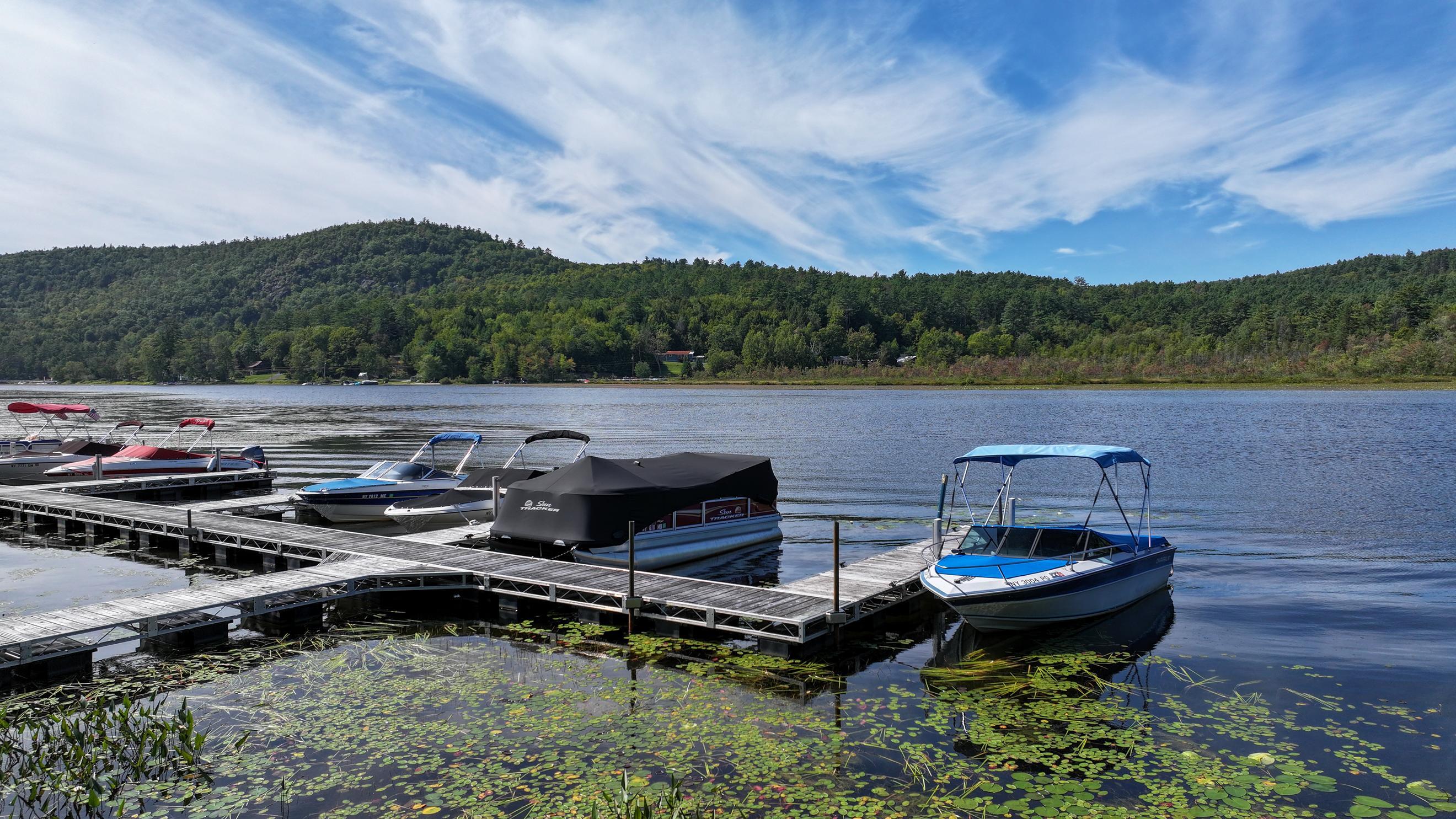 Boats moored at wooden docks on Brant Lake, with lily pads and a forested hillside beyond.