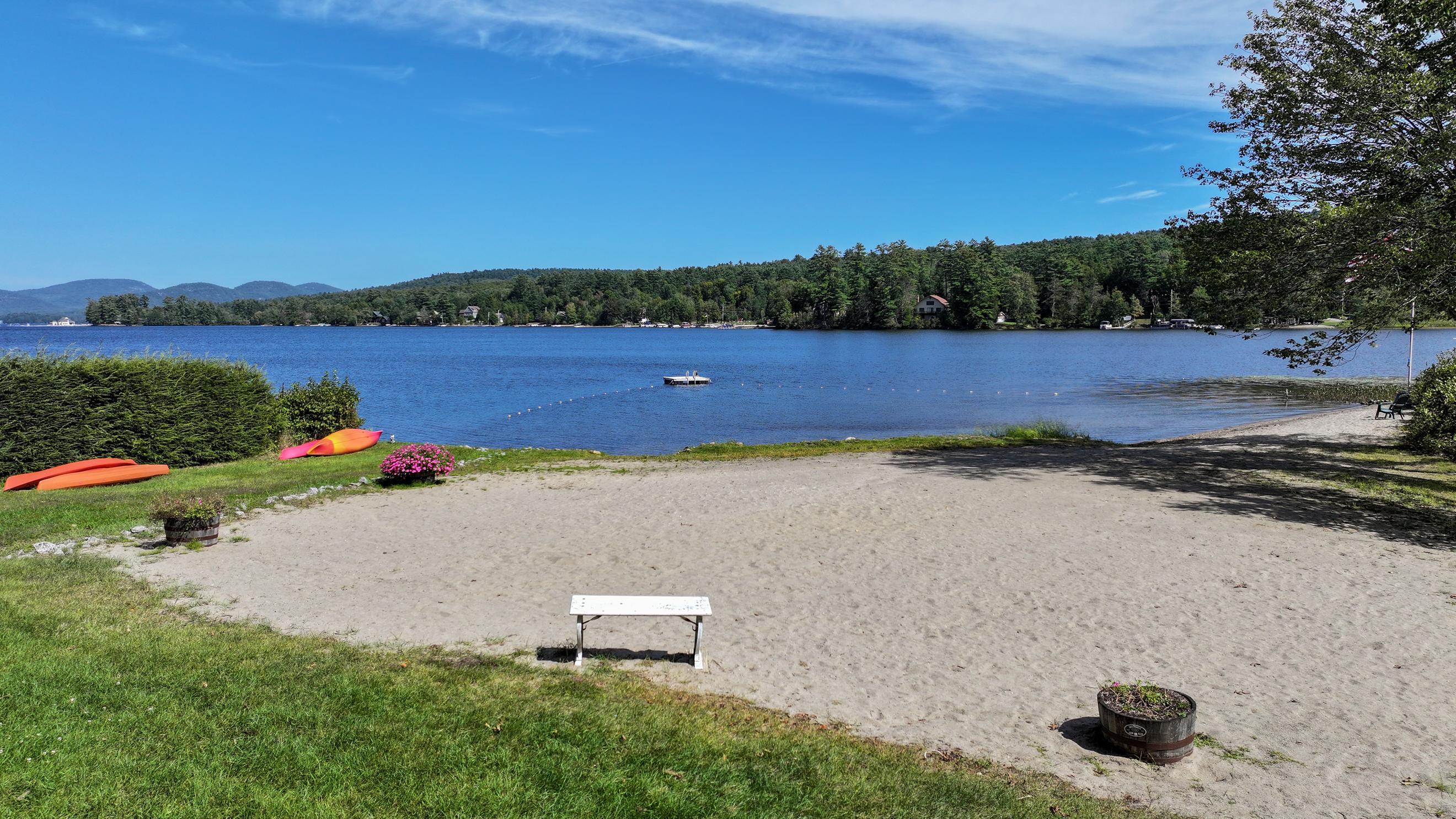 Sandy lakeside beach with a bench and kayaks beside blue water and wooded hills.