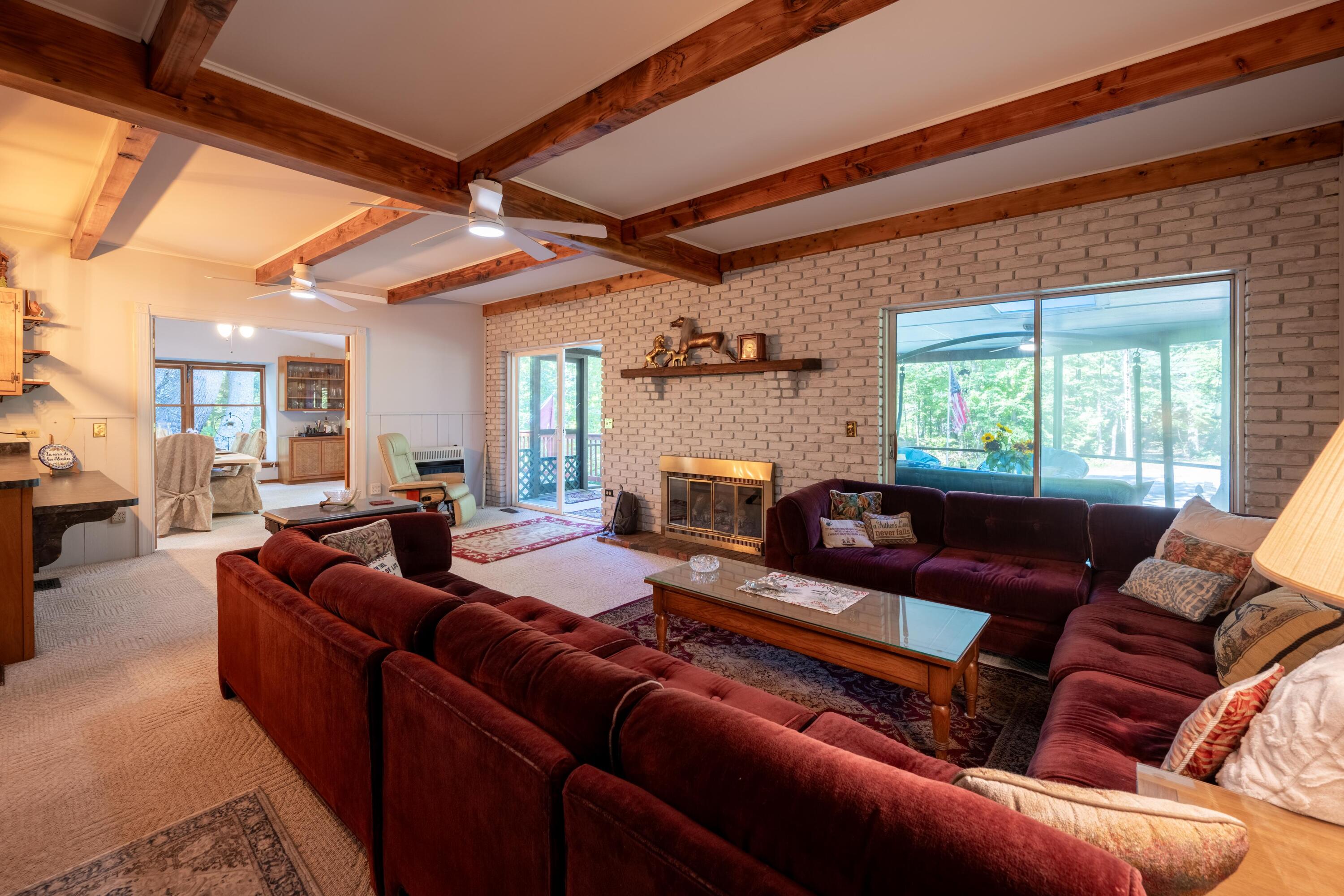 Living room with burgundy sectional sofas, a brick fireplace, exposed wood beams, and large windows.