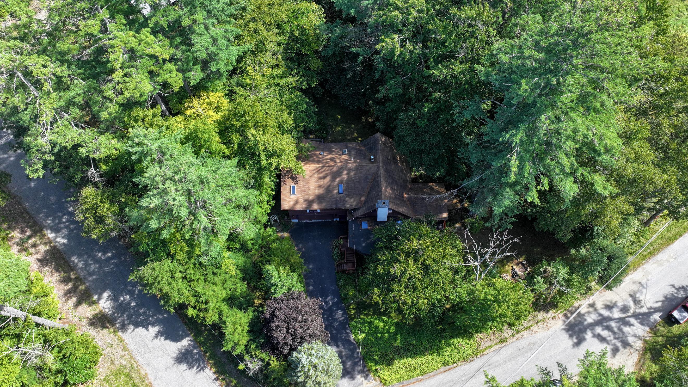 Aerial view of a brown-roofed house nestled among dense trees, with a driveway and roads nearby.