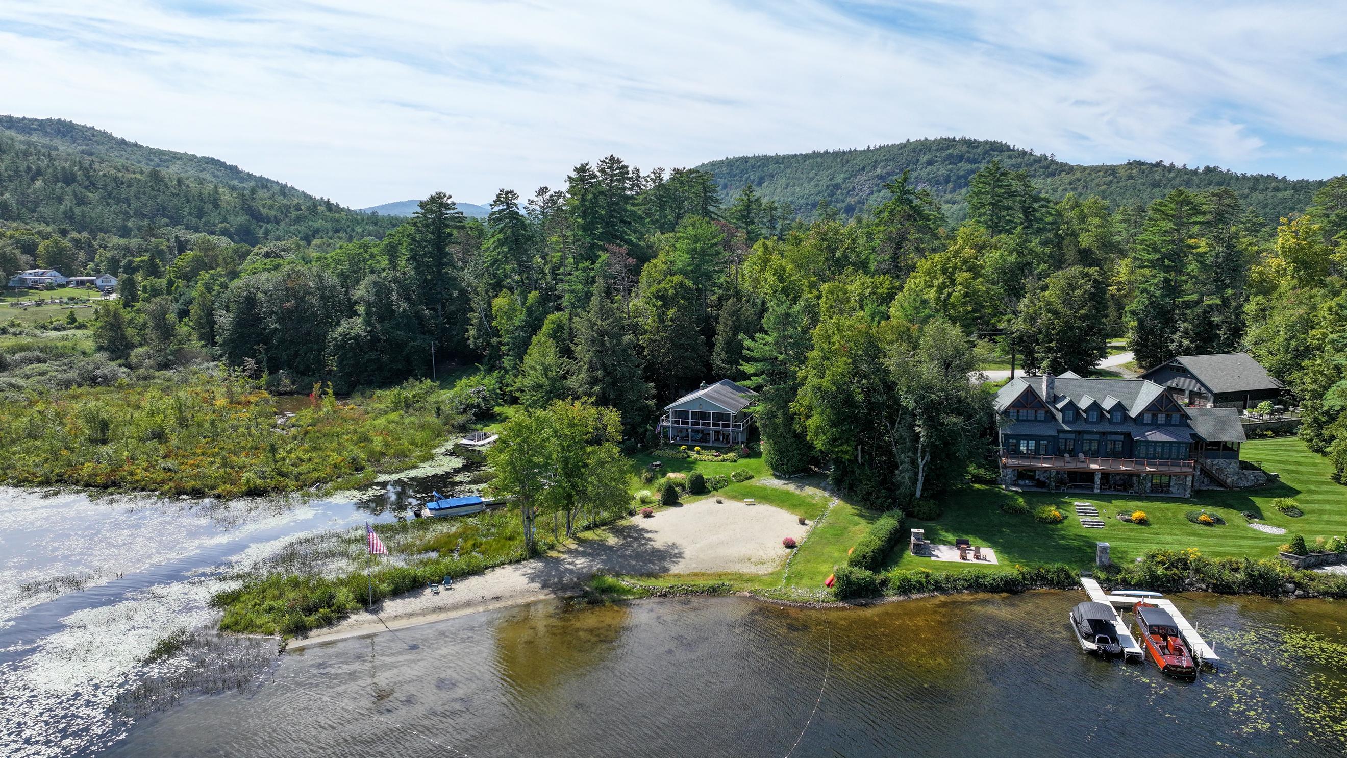 Lakeside home with a sandy beach, two docks, and boats, backed by forested hills.