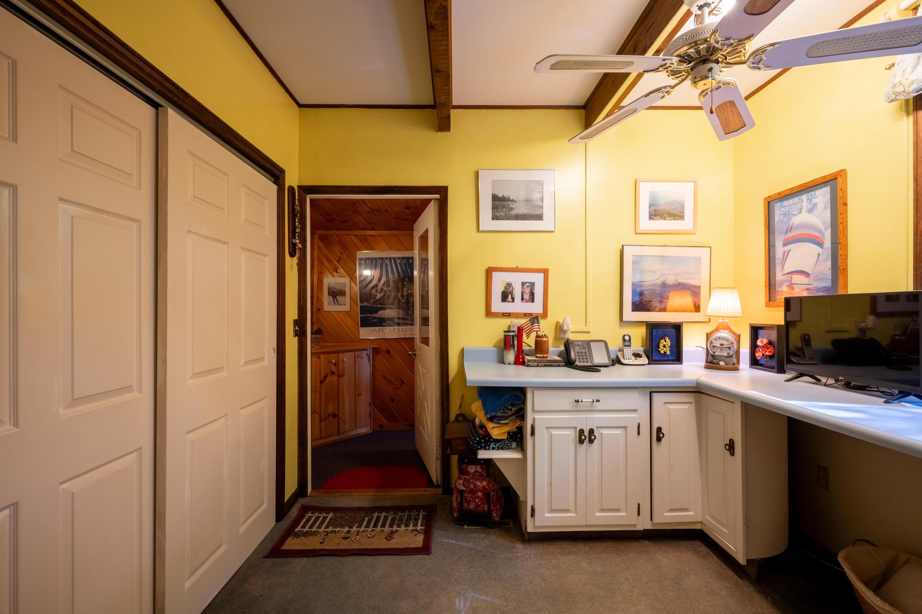 Home office with a white built-in desk, yellow walls, and a doorway to a wood-paneled room.