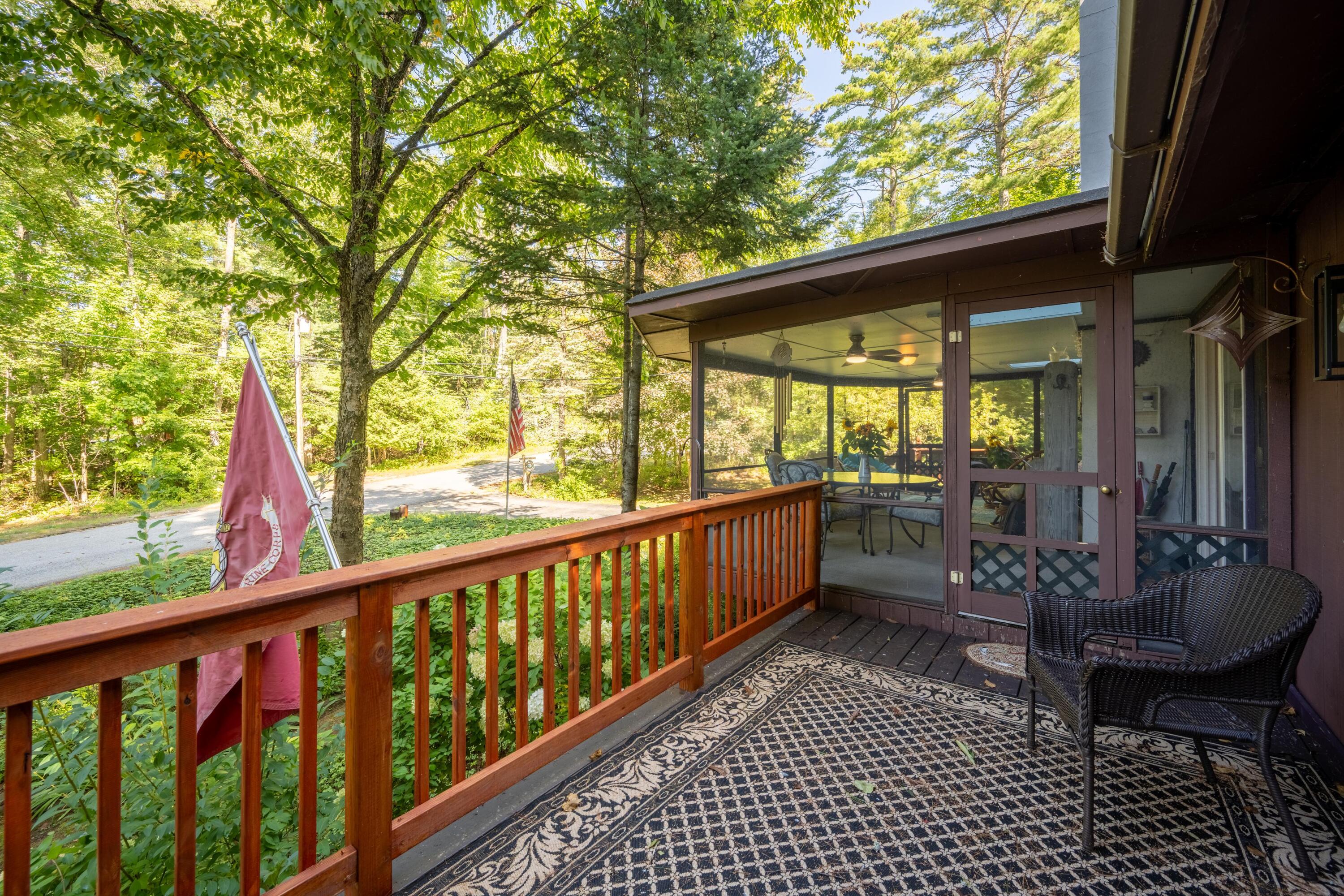 Wooden porch with a patterned rug and wicker chair beside a glass-enclosed sunroom, surrounded by trees.