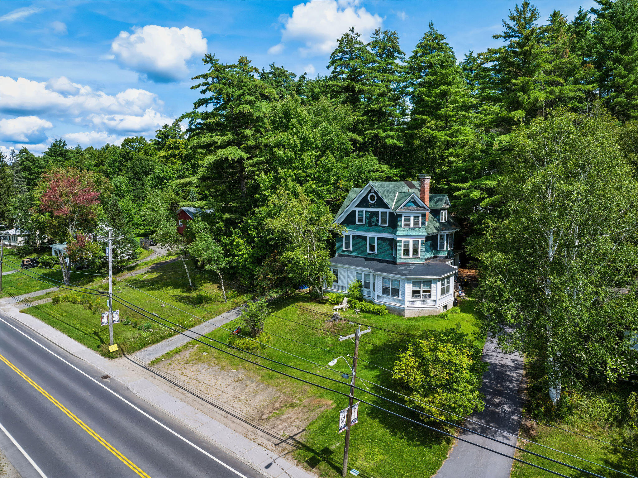Aerial view of a teal-and-white house with a wraparound porch amid mature trees beside a road.