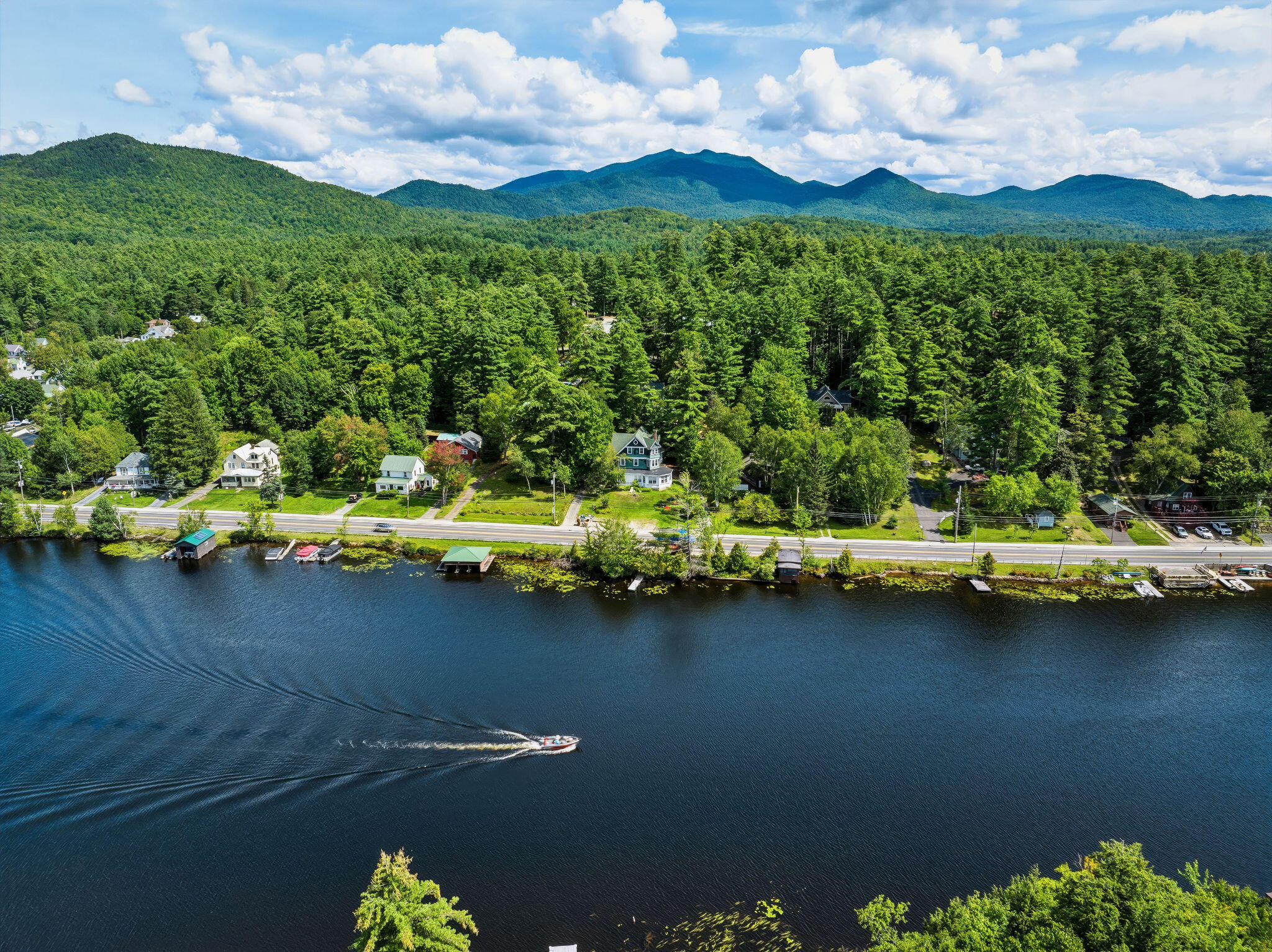 Aerial view of lakeside homes and docks, with a boat on the water and forested mountains beyond.