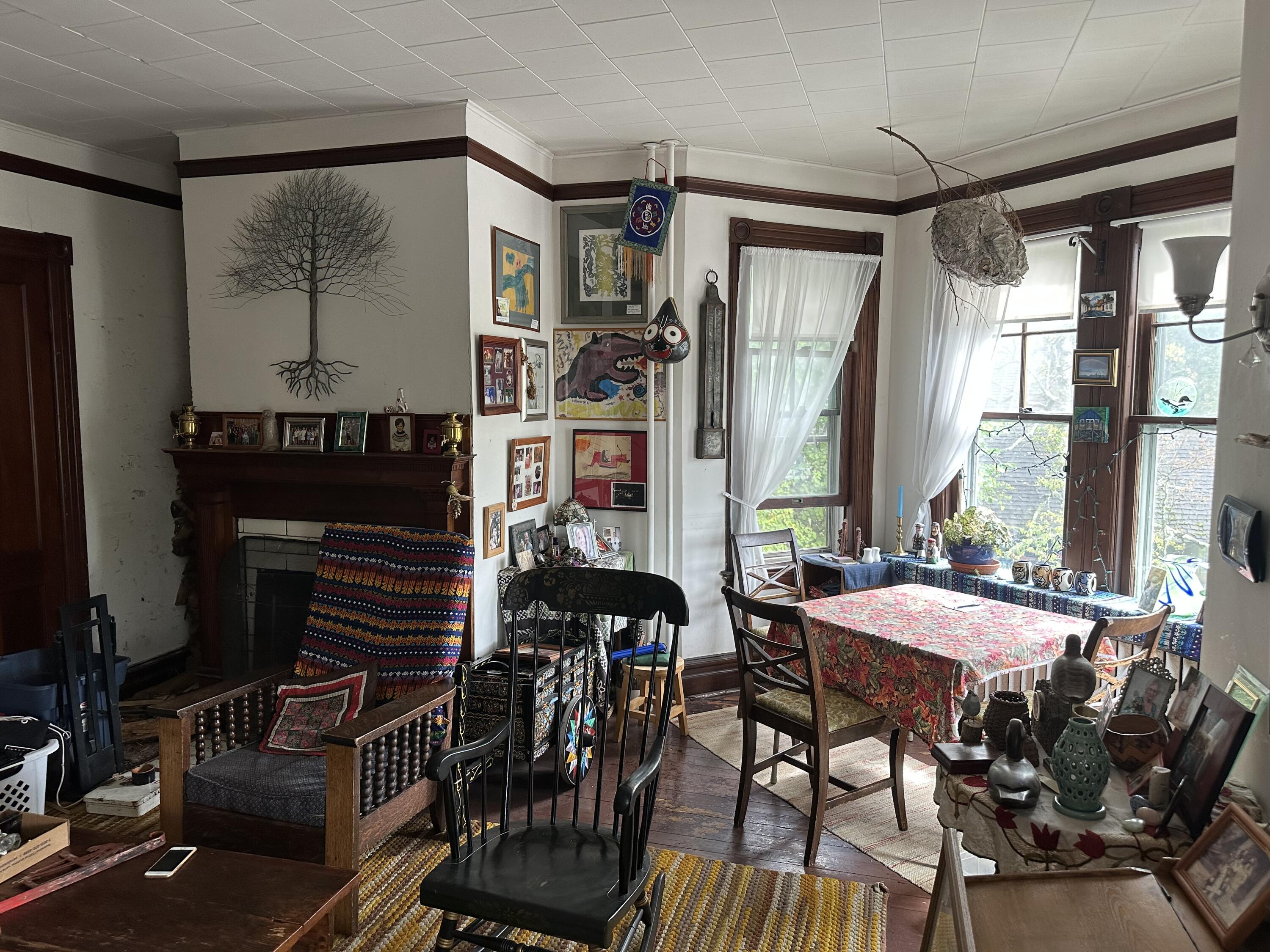 Living and dining room with a fireplace, eclectic artwork, and a floral-covered table by tall windows.