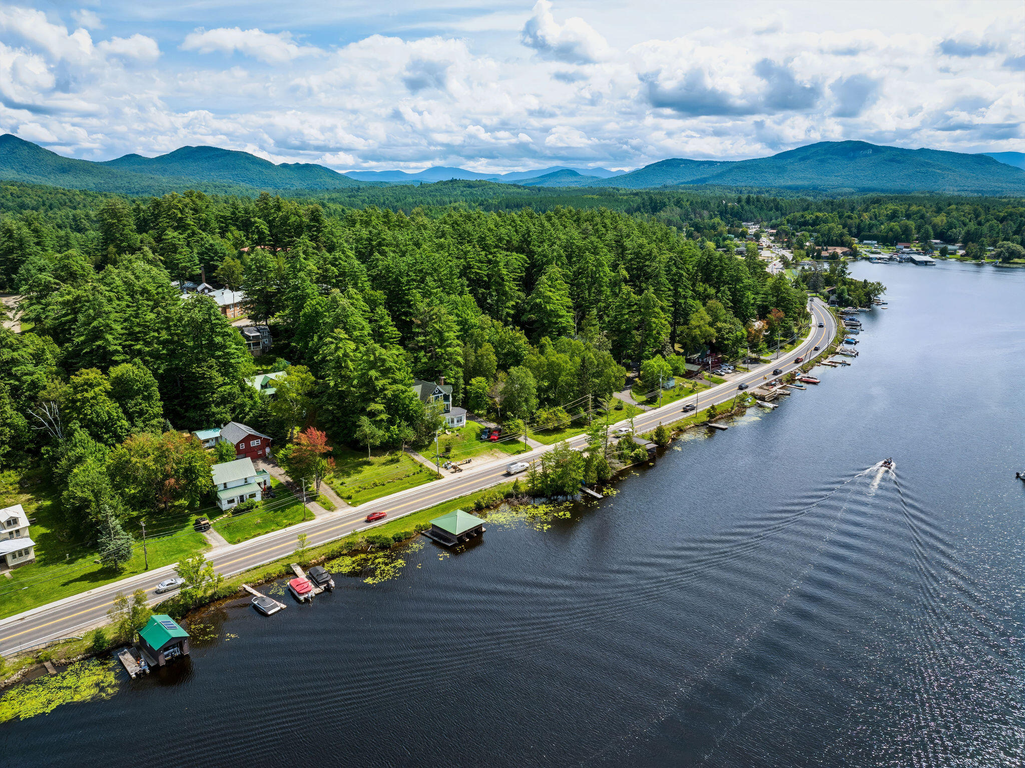 Aerial view of homes along a lake road, surrounded by forest with mountains in the distance.