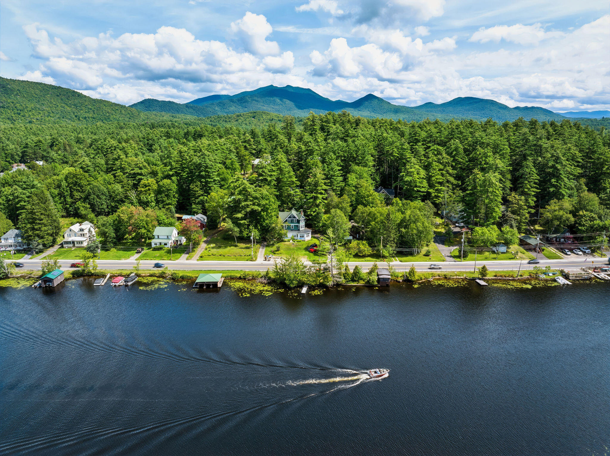 Aerial view of lakeside homes, docks, and a boat on Lake Flower, backed by forested mountains.