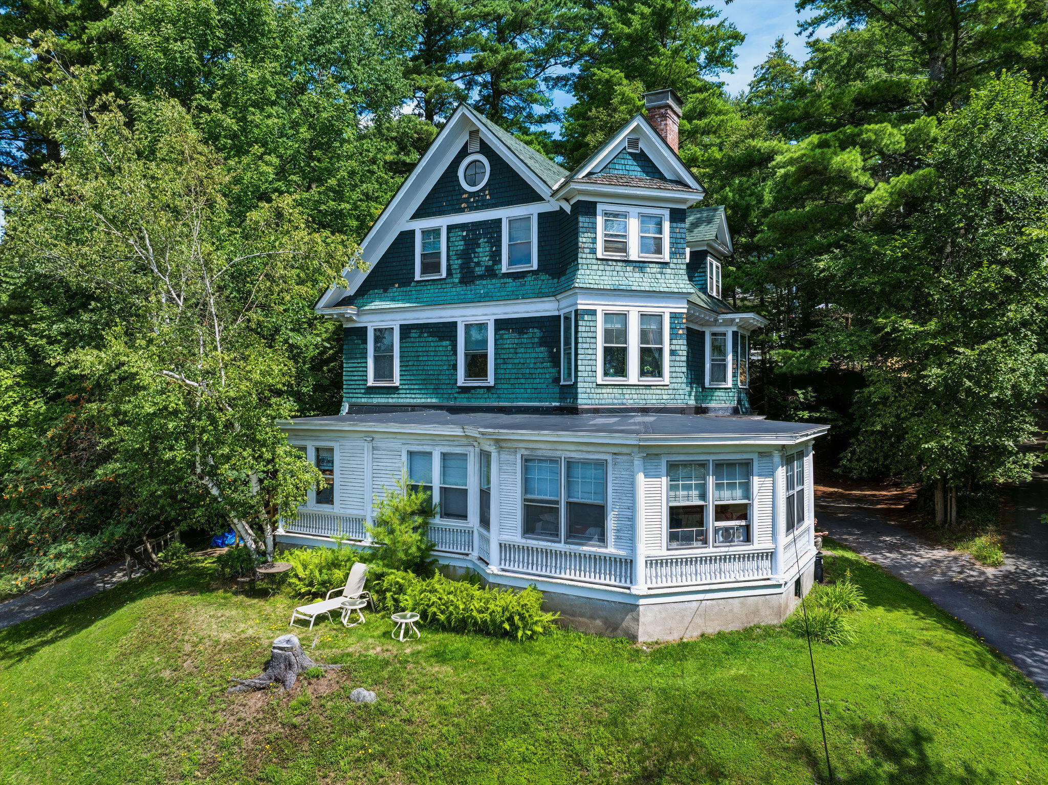 Teal-shingled house with white wraparound porch and steep gabled roof among leafy trees.