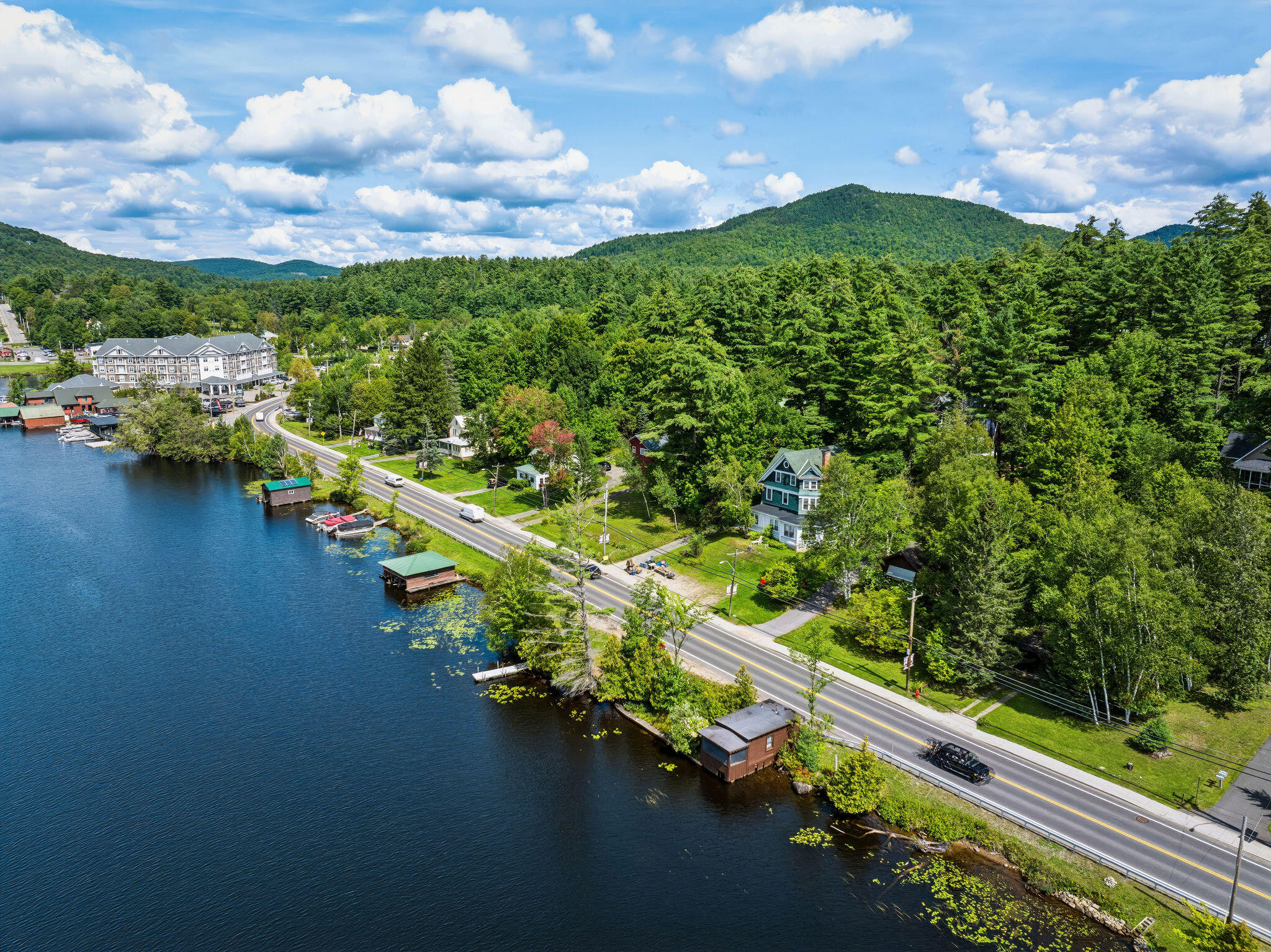 Aerial view of a lakeside home beside a road, with docks, forest, and wooded mountains.