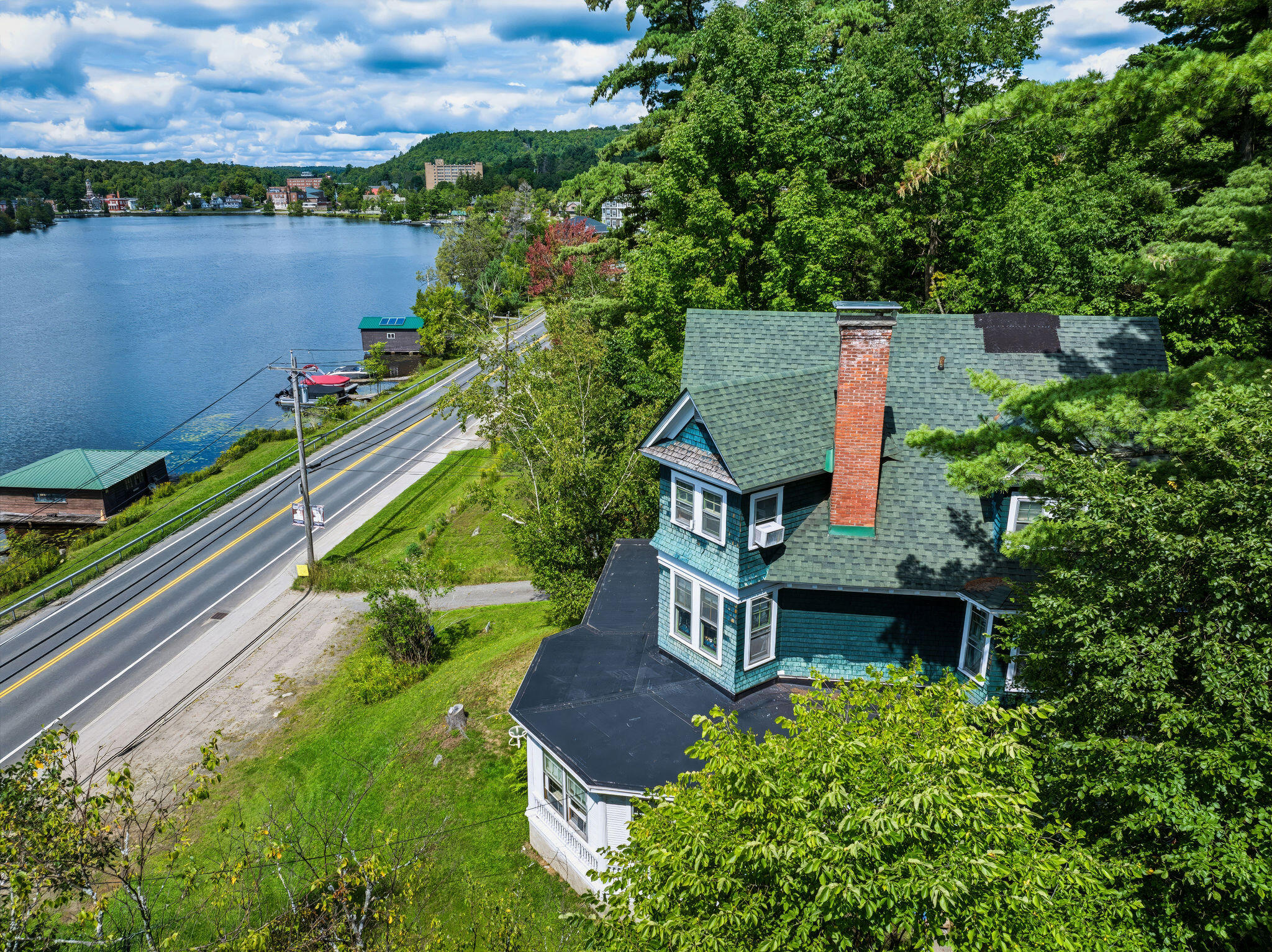 Aerial view of a blue house beside a road, with a lake and wooded hills nearby.