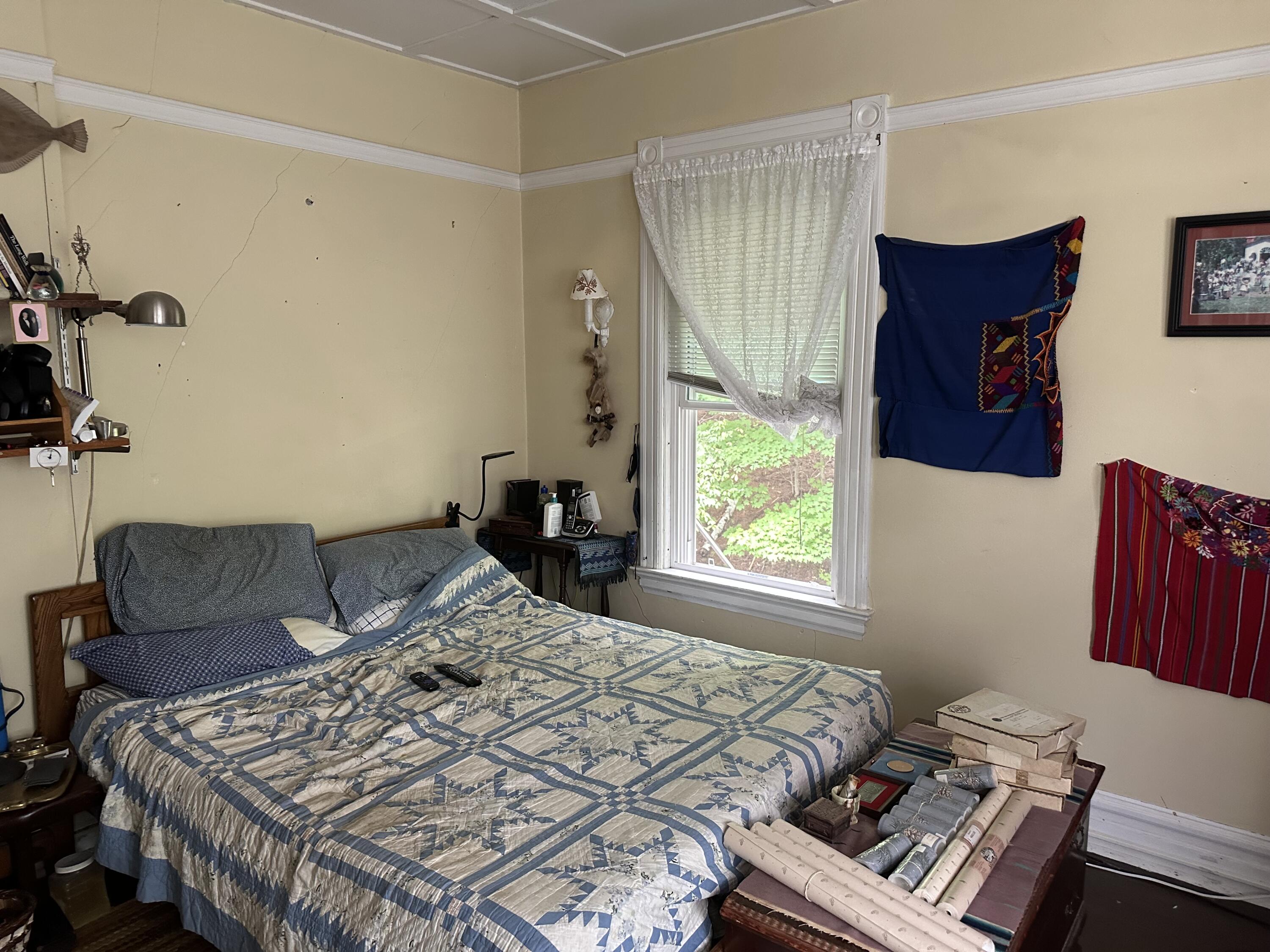 Bedroom with a geometric quilt-covered bed, lace-curtained window, and colorful wall hangings.