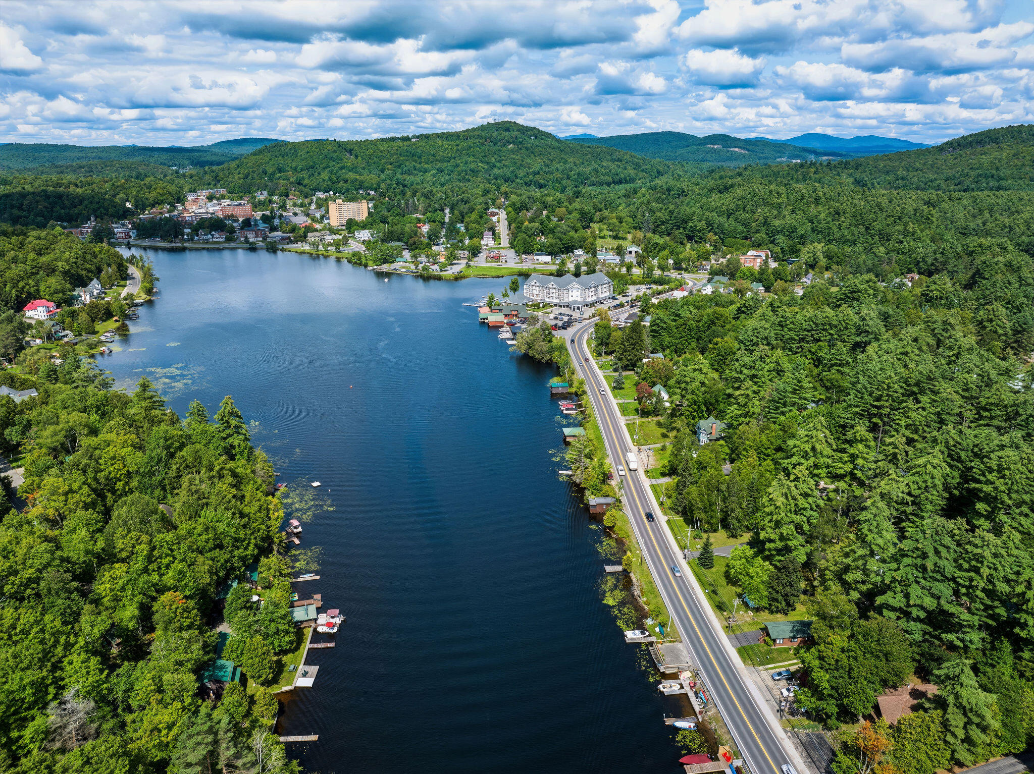 Aerial view of a forested lakeside town, with a road following the water and mountains beyond.