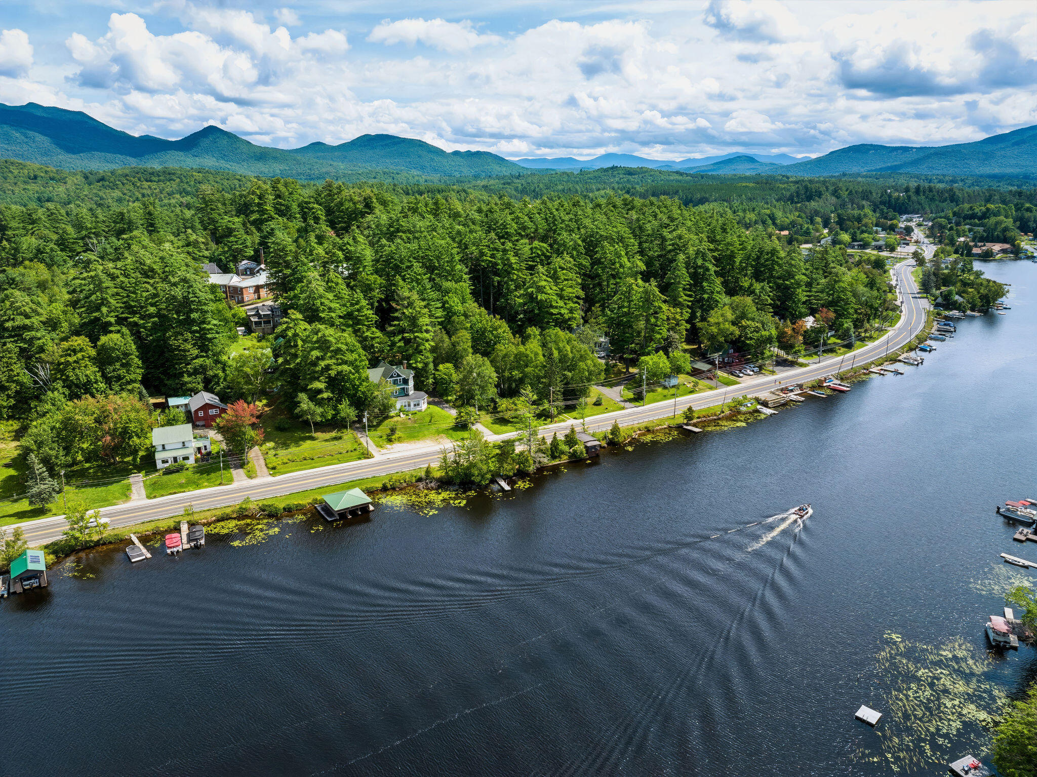 Aerial view of a lakeshore road, wooded homes, docks, and boats with mountains beyond.