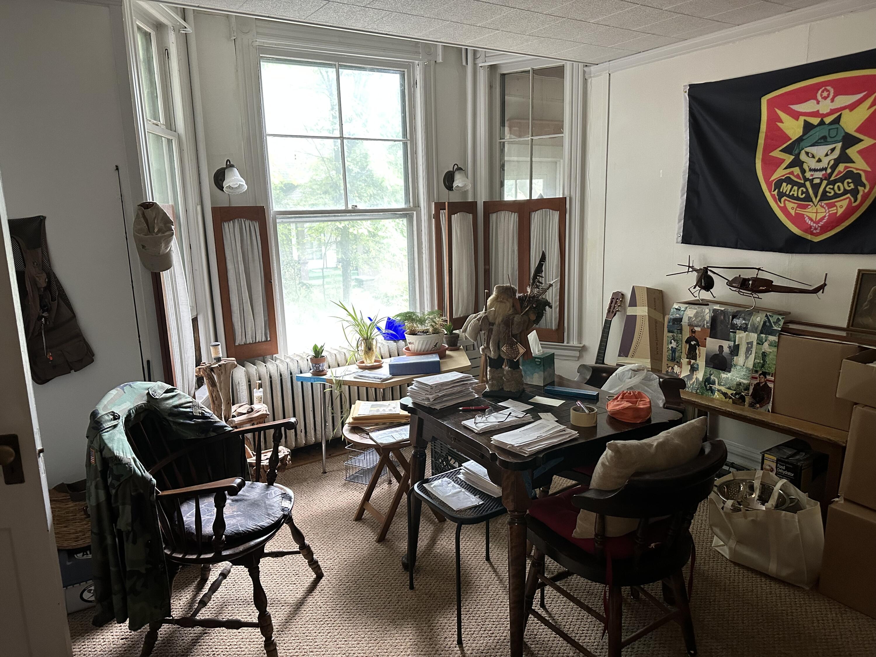 Cluttered office with a large window, paper-covered desk, plants, and a MAC V SOG flag.