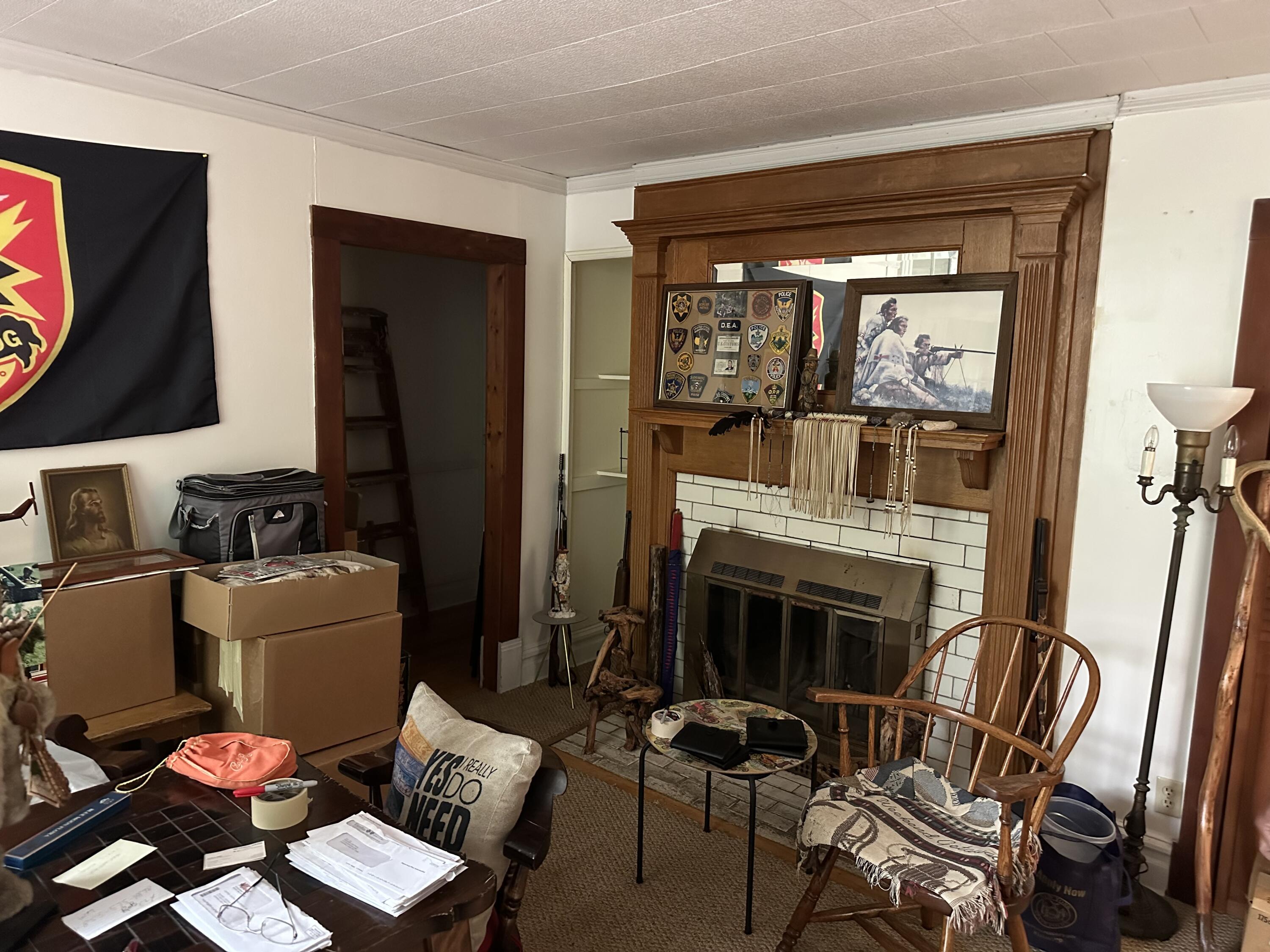 Cluttered living room with a dark wood fireplace, white tile, and boxes and belongings throughout.