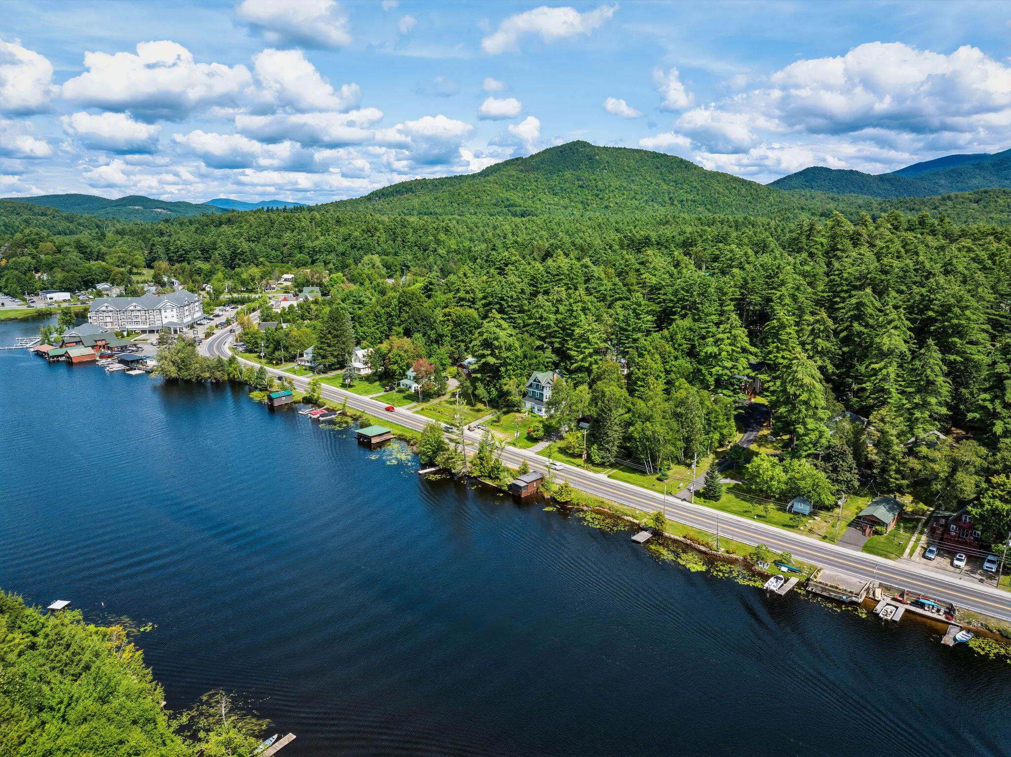 Aerial view of a lakeside road and homes, with forested Adirondack hills beyond.