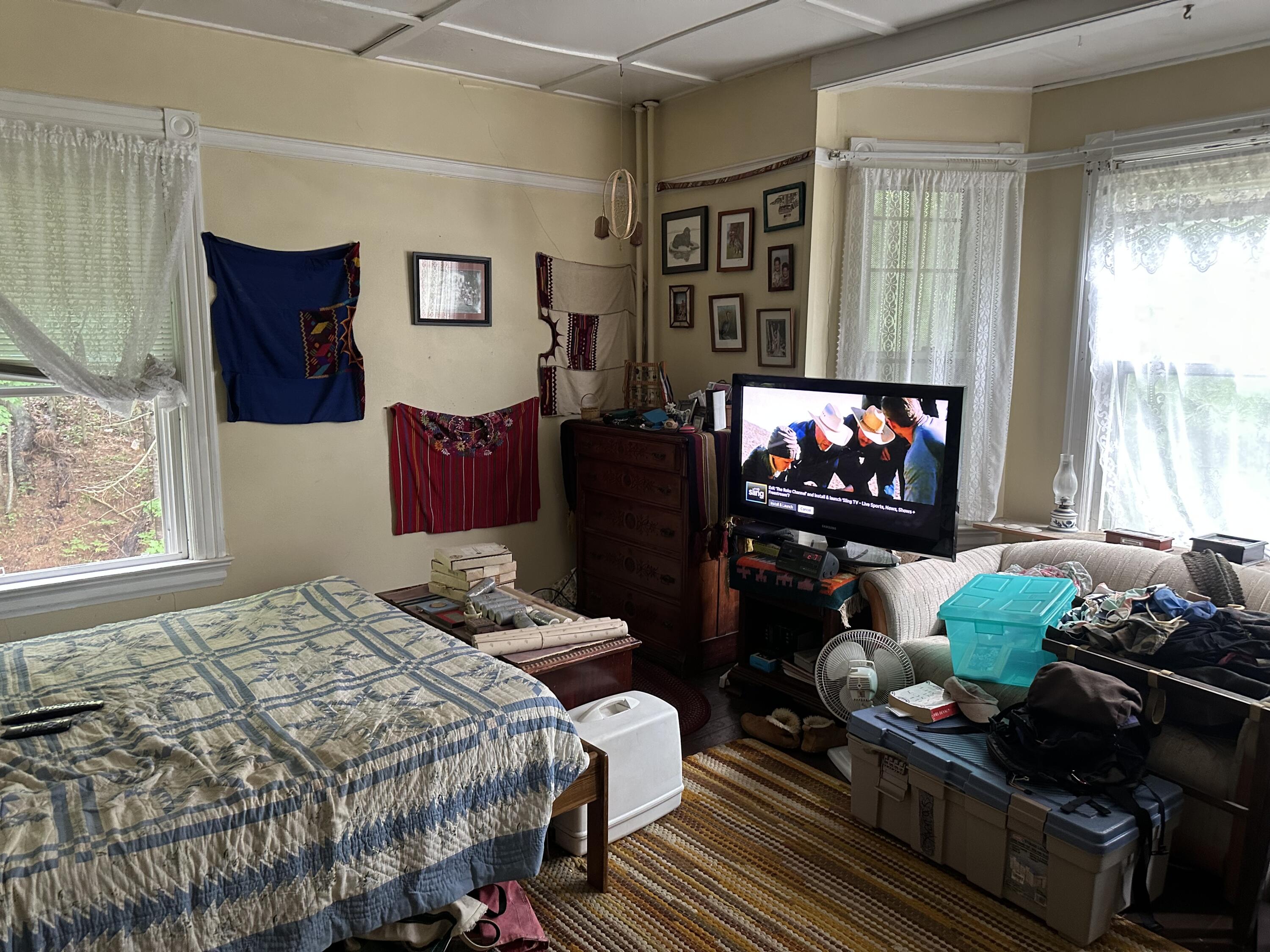 Cluttered bedroom with a blue-and-beige quilted bed, lace-curtained windows, and a television.