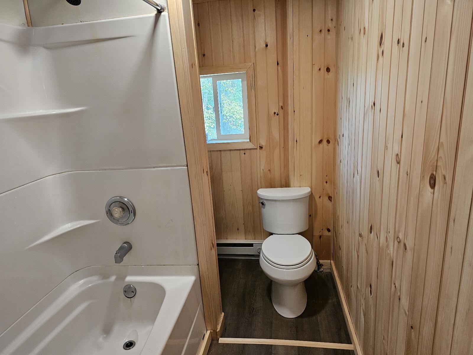 Bathroom with a white tub and shower, toilet, and natural wood-paneled walls.