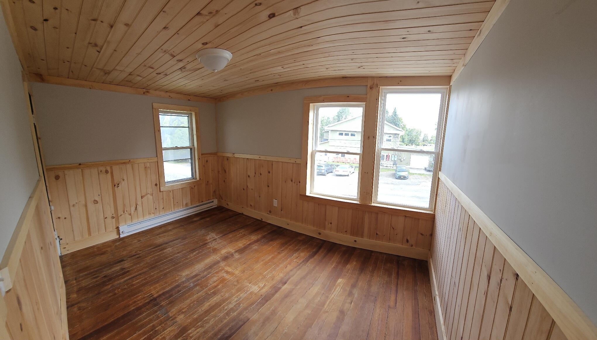 Empty room with a wood-plank ceiling, wood wainscoting, hardwood floors, and windows.