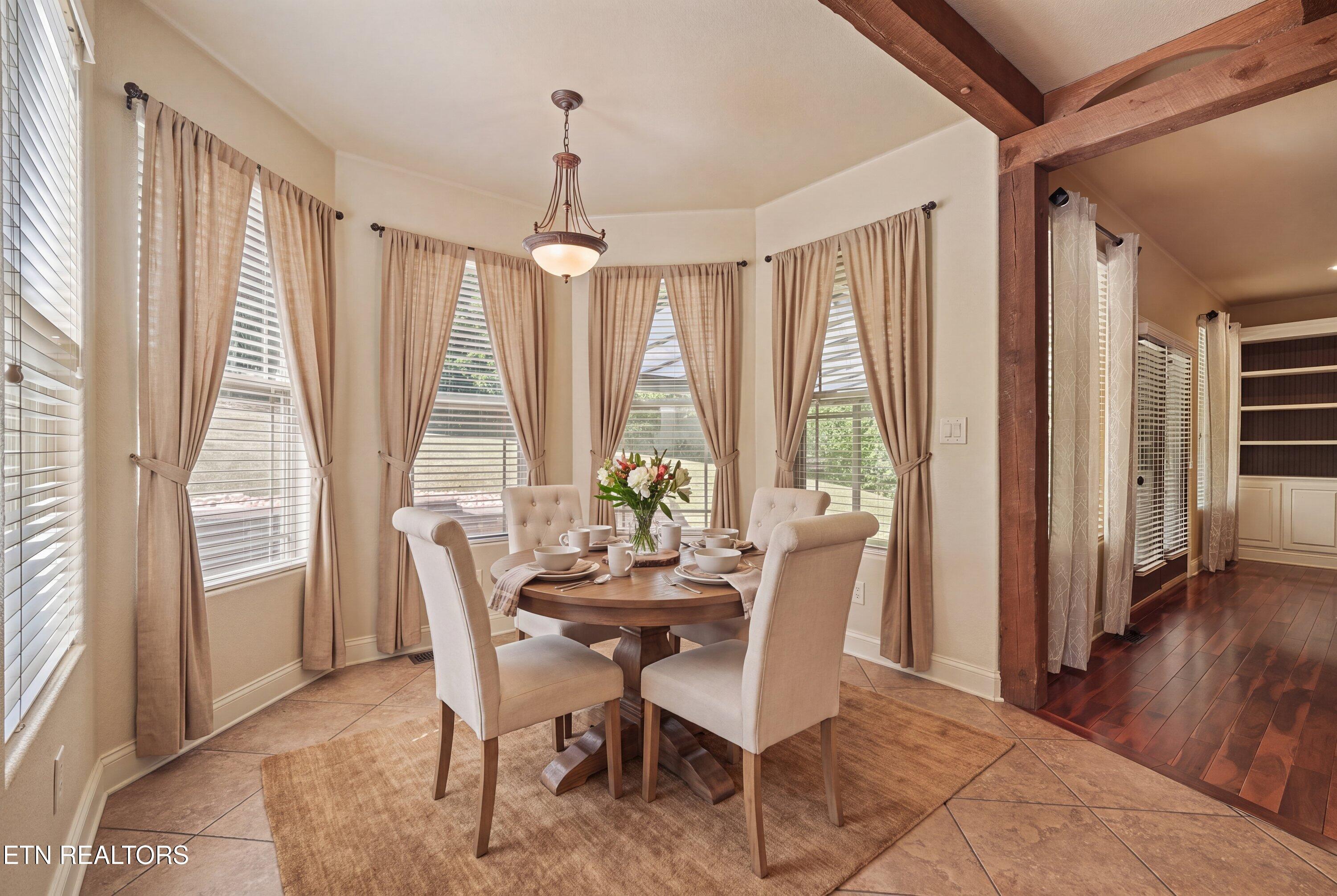 Dining room with a set round table, tall curtained windows, and exposed wood beams.