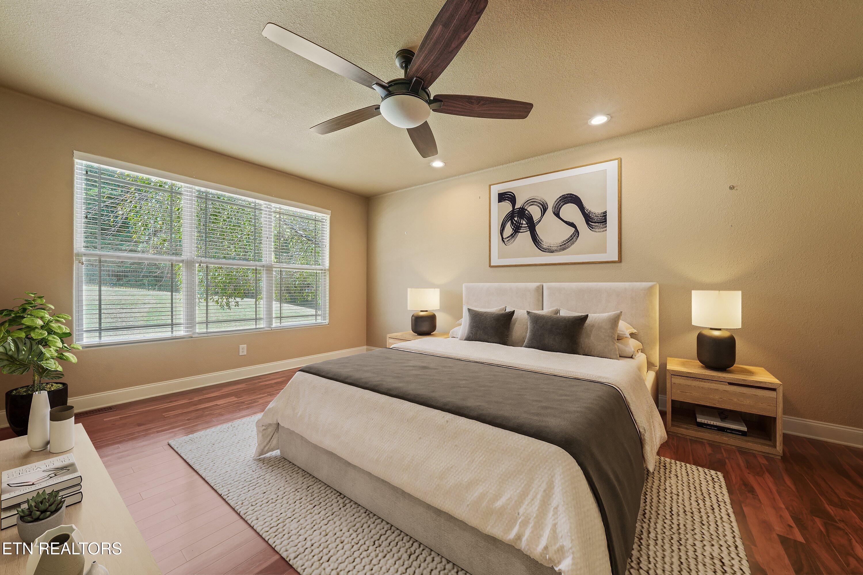 Staged bedroom with a large cream-and-charcoal bed, wide window, hardwood floors, and ceiling fan.