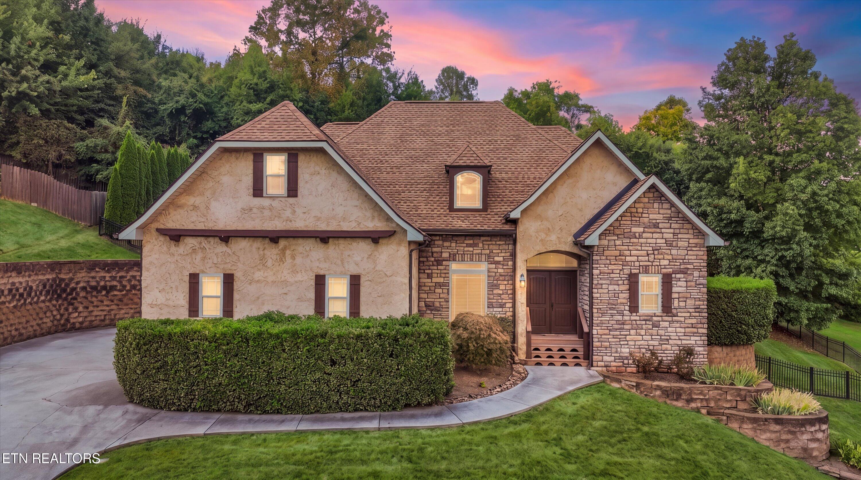 Two-story stone and stucco home with trimmed hedges and a curved front walkway at sunset.