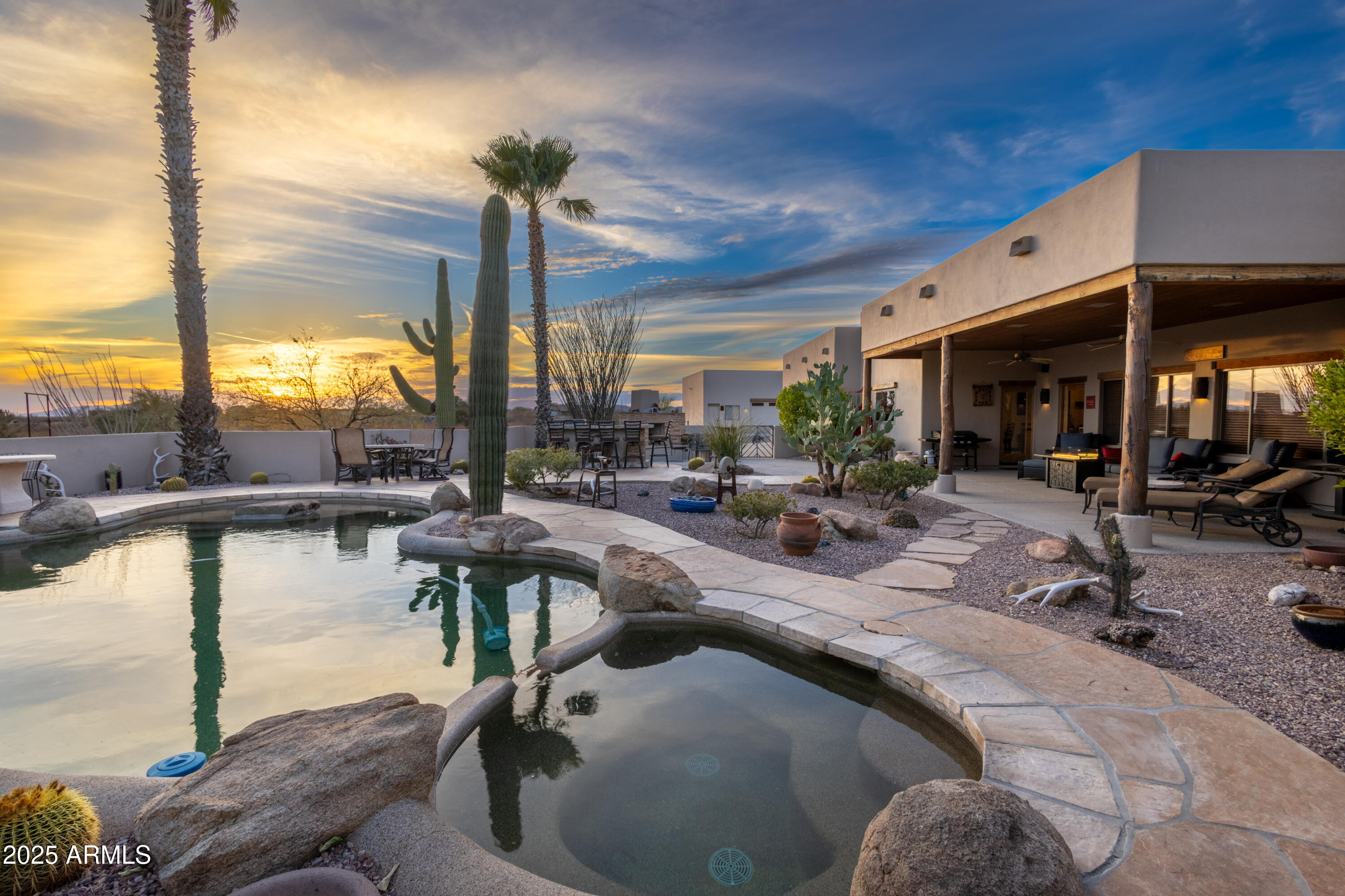 Desert home backyard with a curved pool, covered patio, and saguaro cactus at sunset.