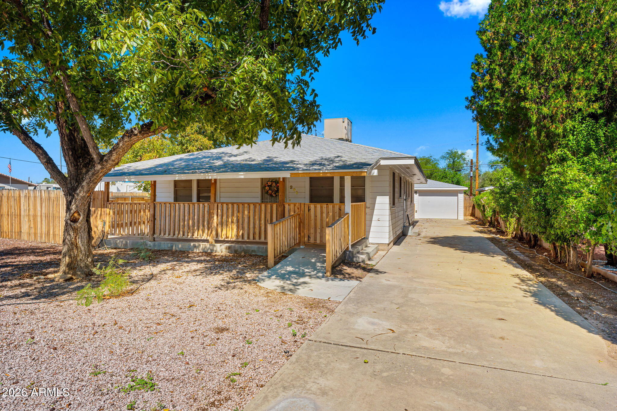 Single-story house with a covered wooden-railed porch, mature shade trees, and a driveway to the garage.