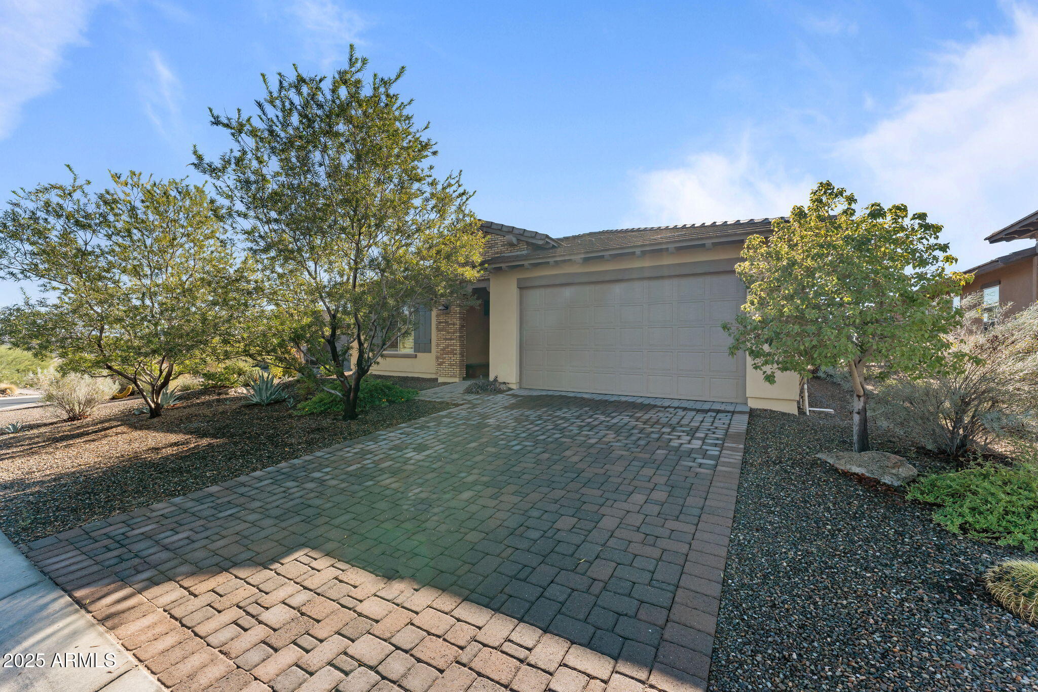 Home exterior with a two-car garage, wide paver driveway, and mature trees.