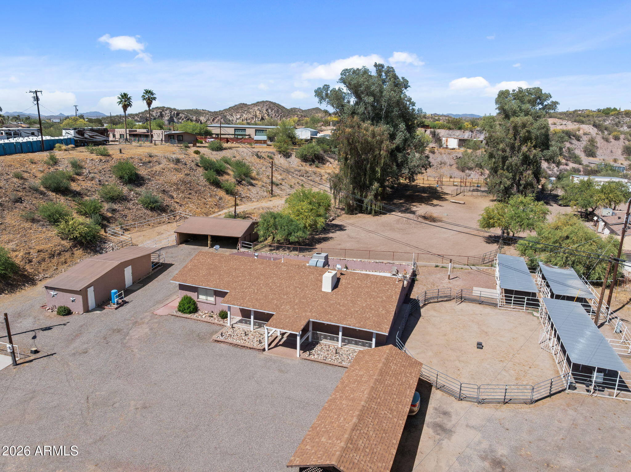 Aerial view of a desert property with a home, outbuildings, fenced pens, and covered horse stalls.