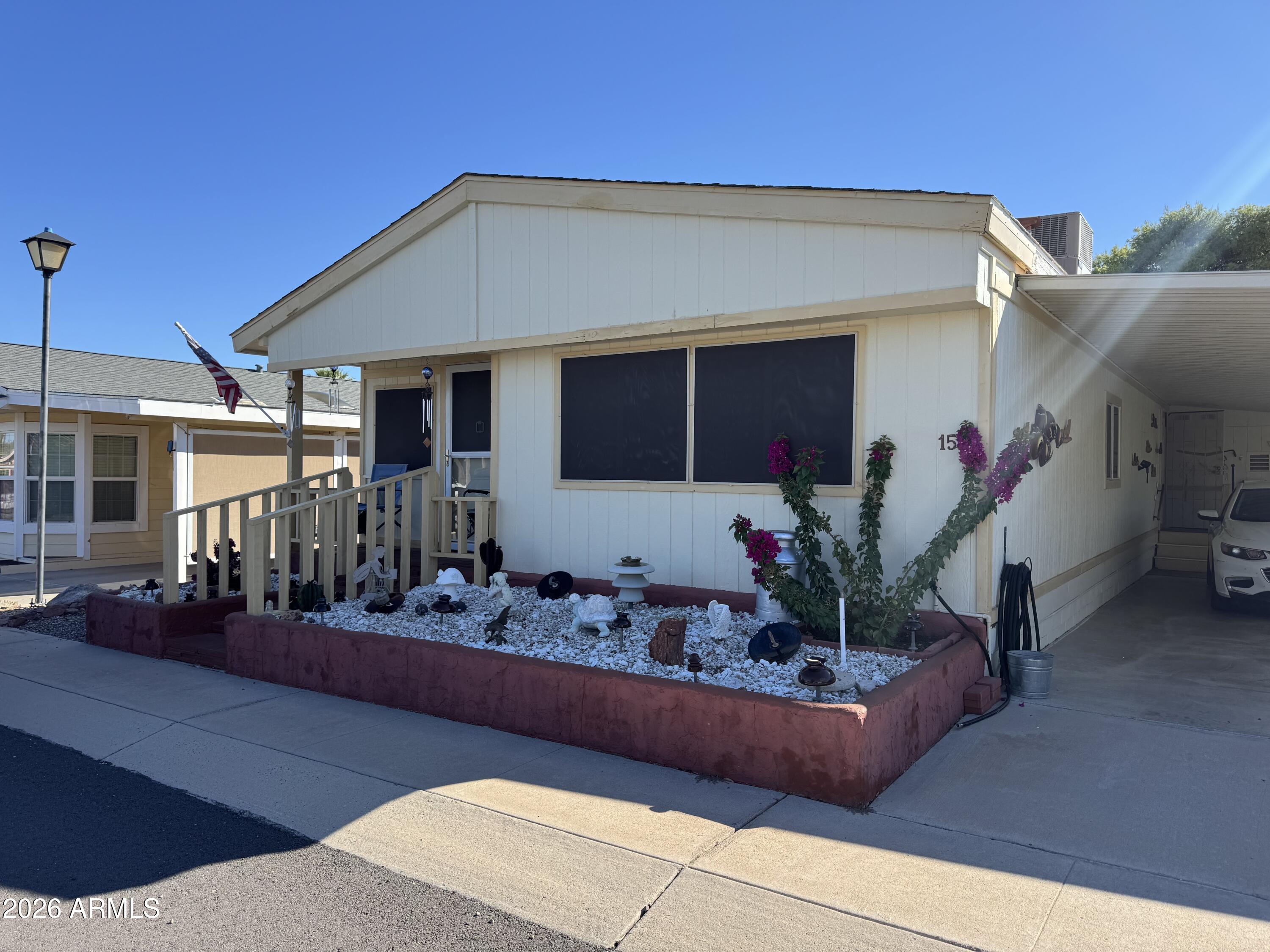 White manufactured home with a raised porch, gravel garden, flowering shrubs, and covered driveway.
