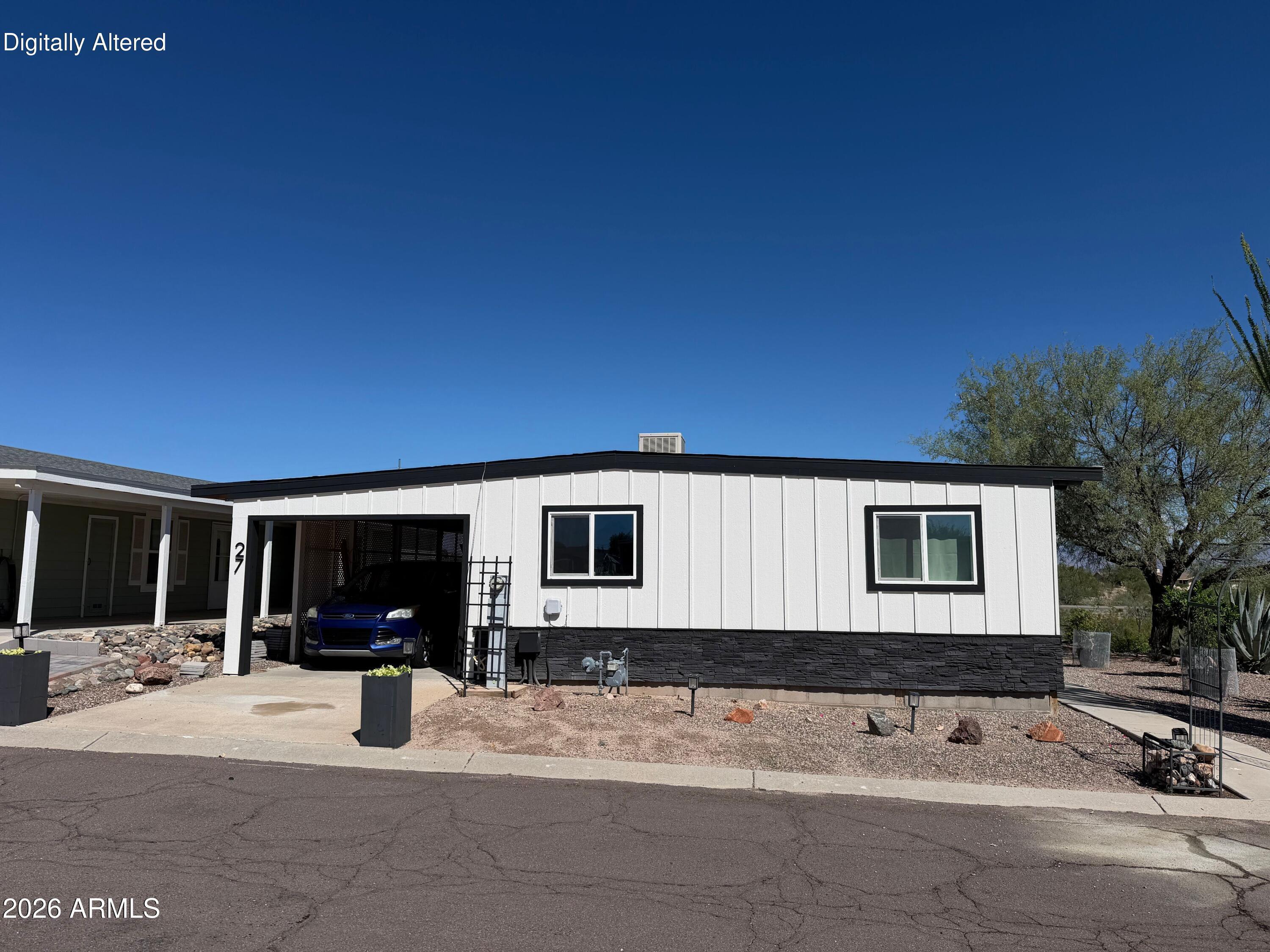 White manufactured home with black trim, a covered carport, and gravel desert landscaping.