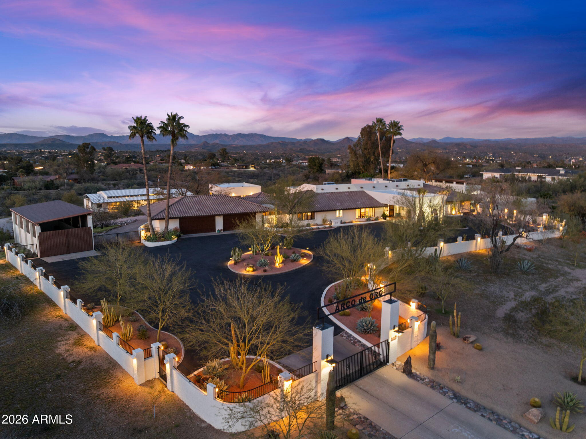 Aerial view of a walled Southwestern property with a gated entrance, desert landscaping, and distant mountains at dusk.