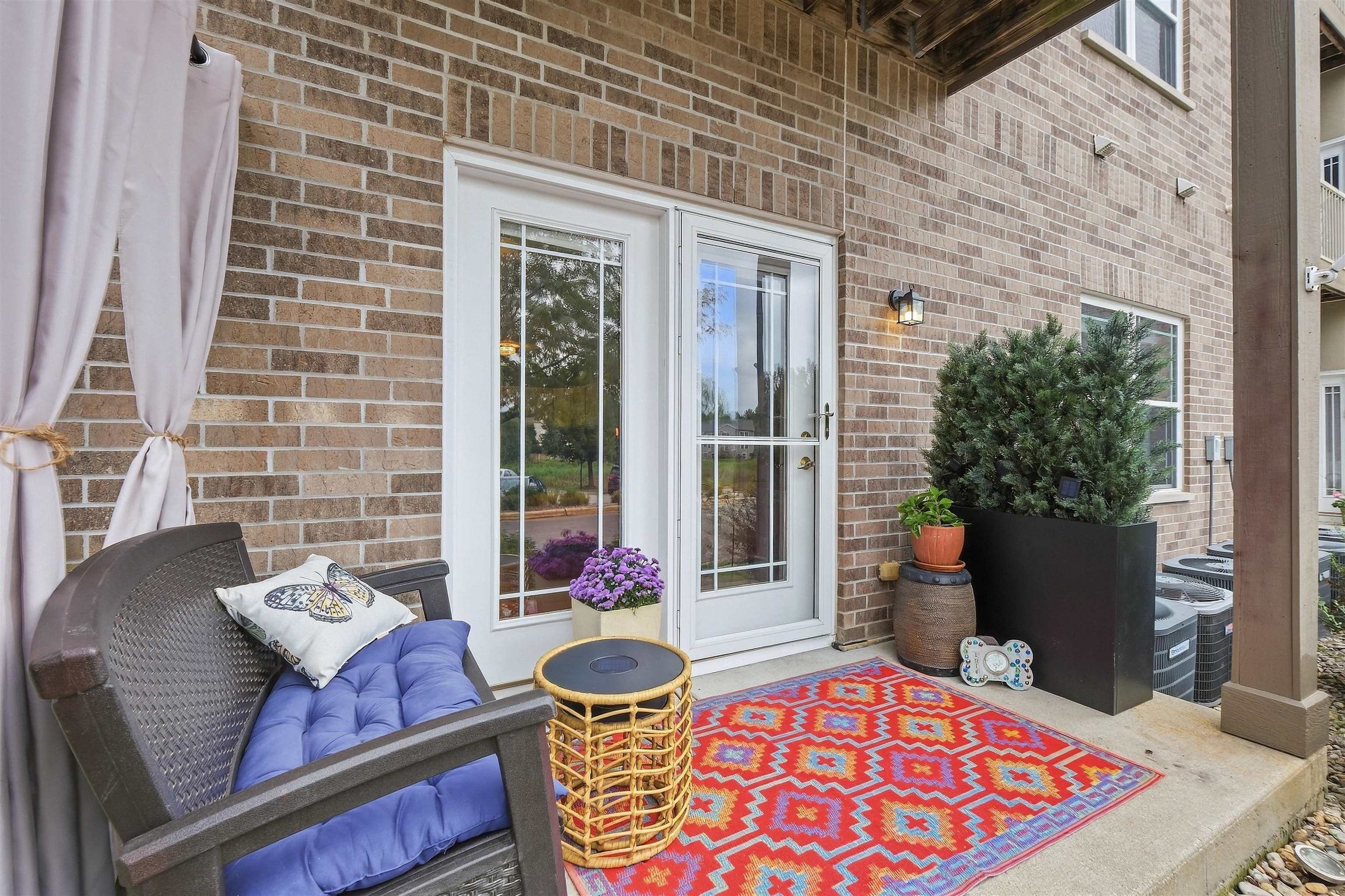 Covered brick patio with a cushioned chair, colorful rug, potted greenery, and glass door.