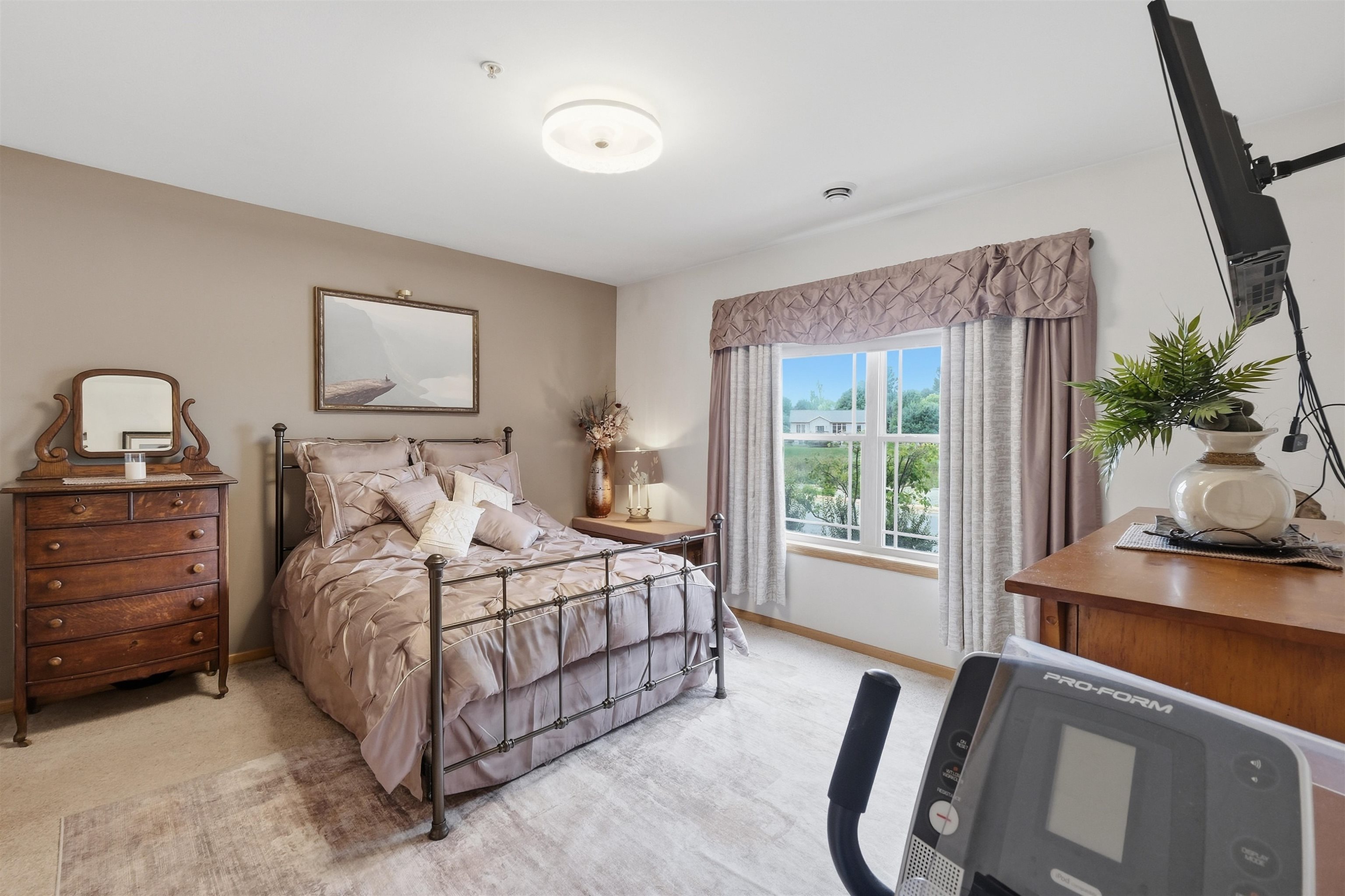 Bedroom with a metal bed, mauve bedding, wooden dresser, and large curtained window.