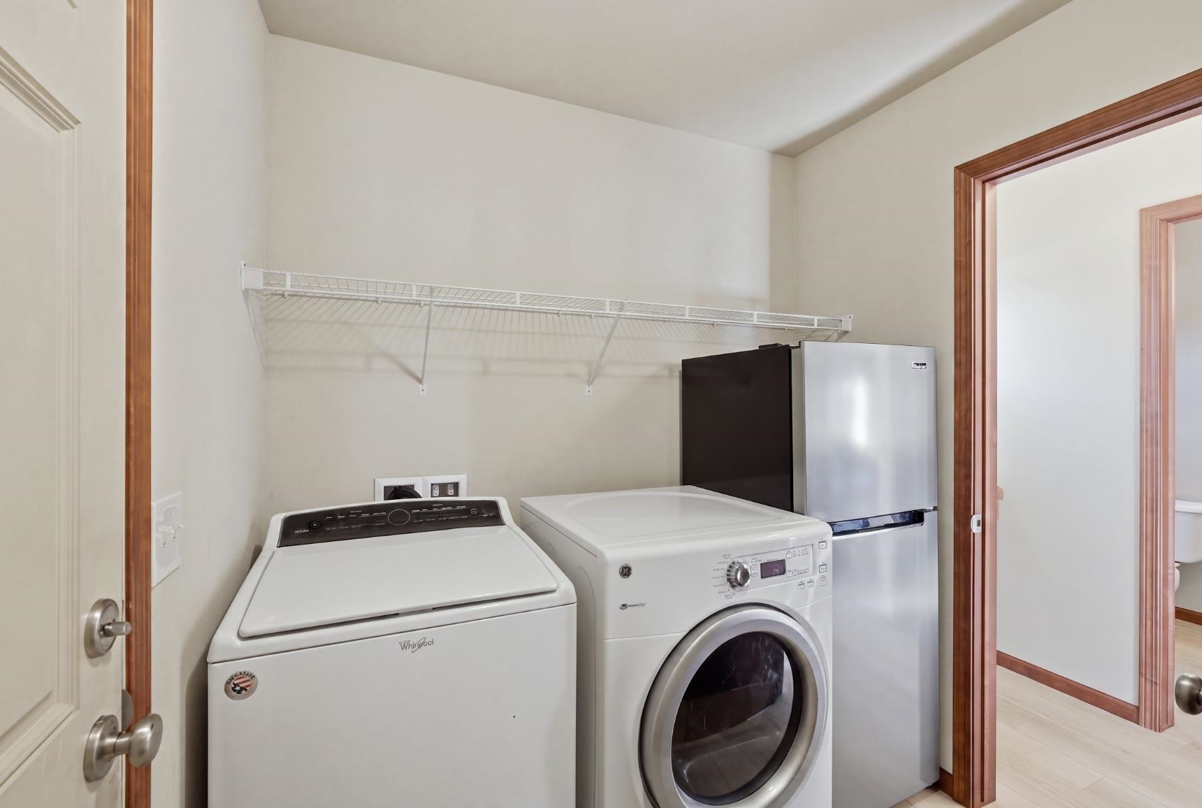 Laundry room with a top-load washer, front-load dryer, refrigerator, and long wire shelf.